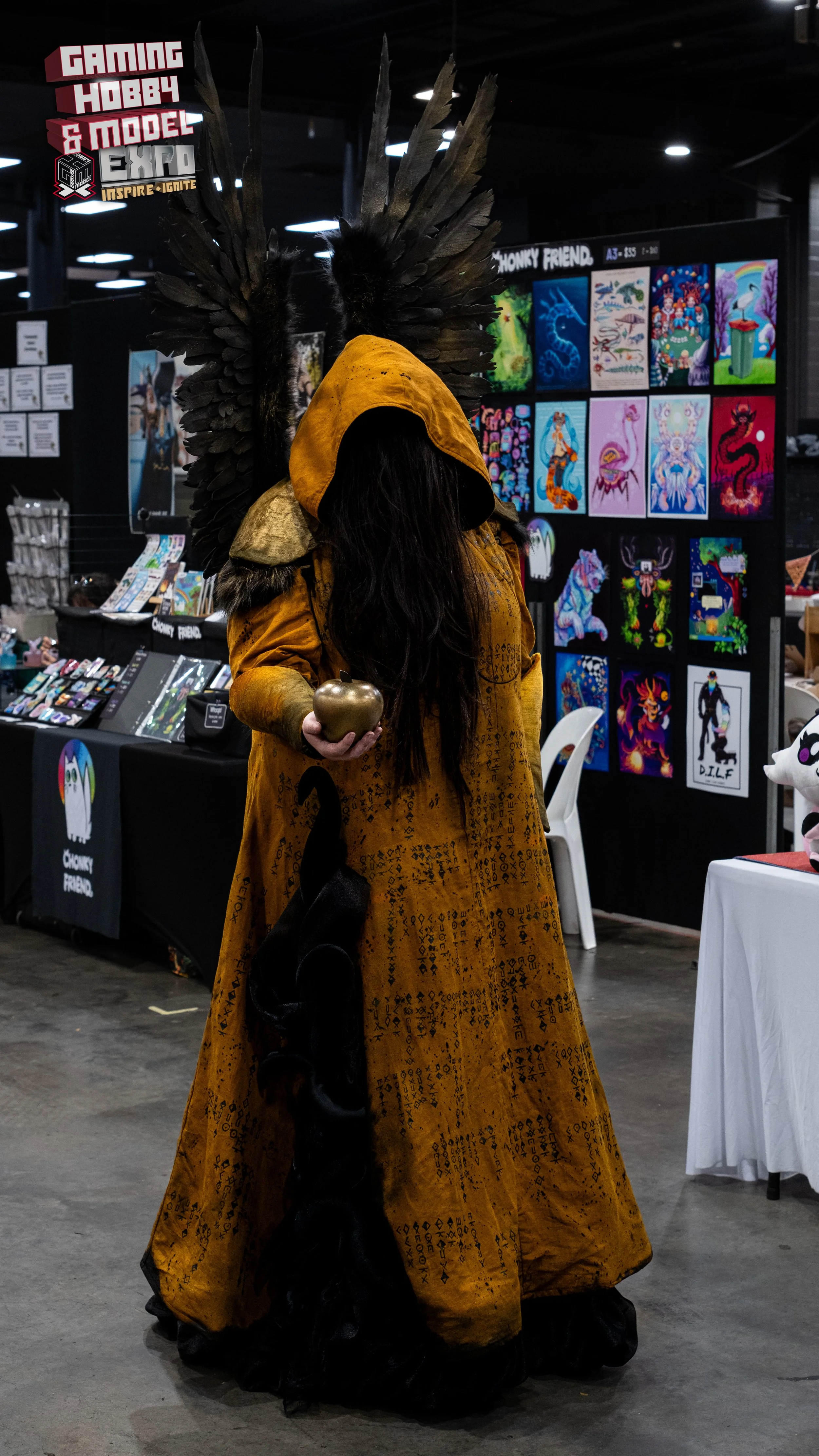 Person dressed in elaborate costume with a large feathered headdress, wearing a long yellow robe, and holding a golden apple, standing at a convention booth with colorful artwork on display.