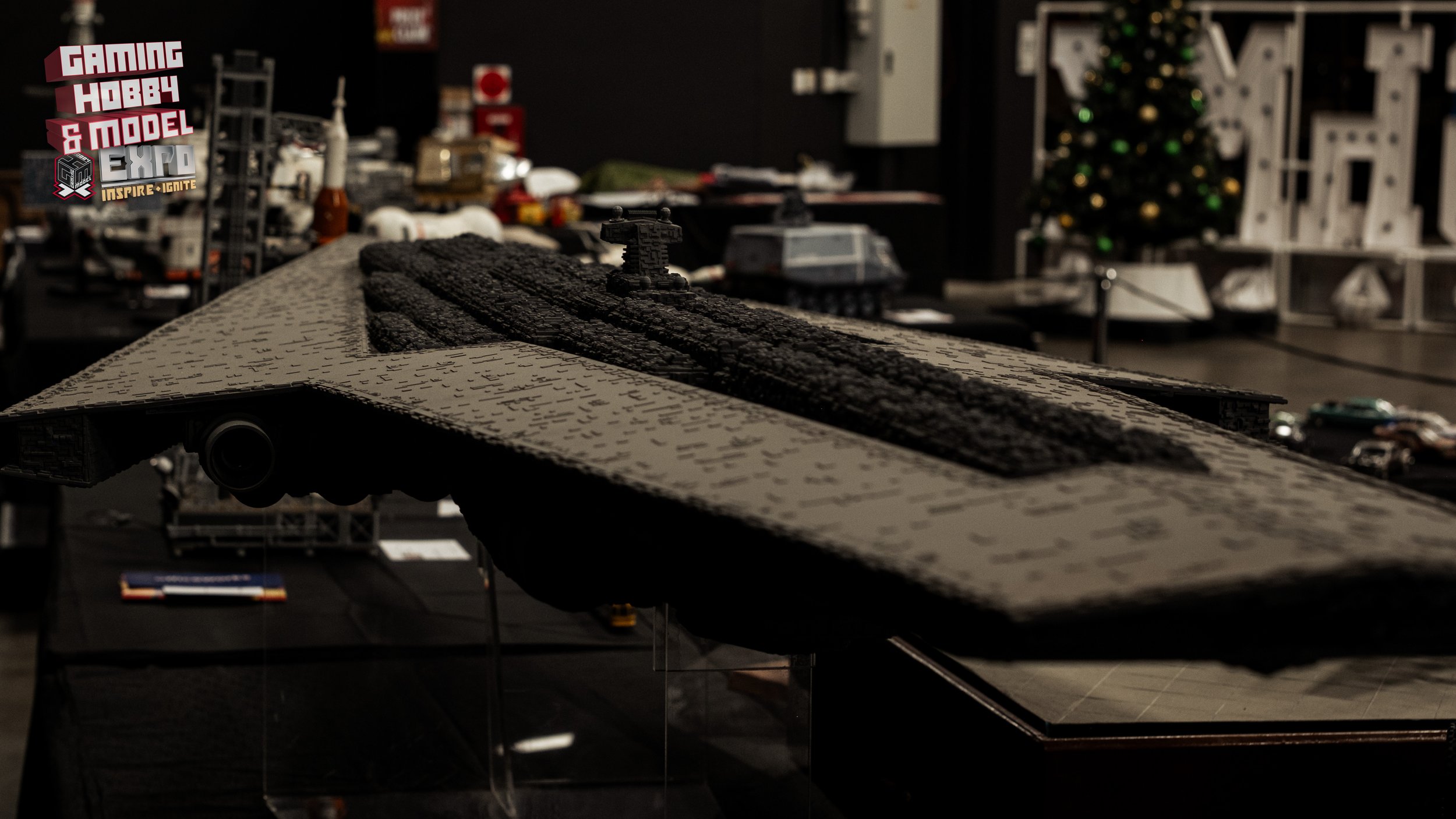 Close-up of a LEGO spaceship model displayed on a black table at an exhibition, with a Christmas tree and shelves with other models in the background.
