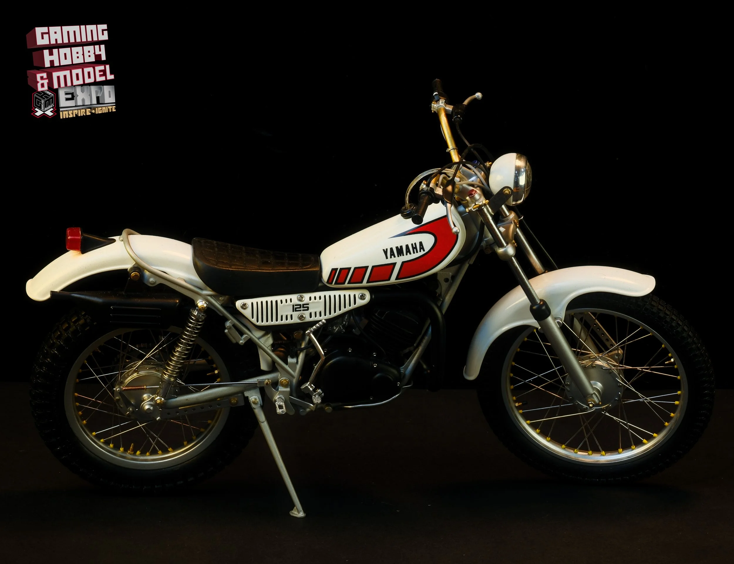 A vintage white Yamaha motorcycle with black seat and red detailing on a black background, displayed at an event.