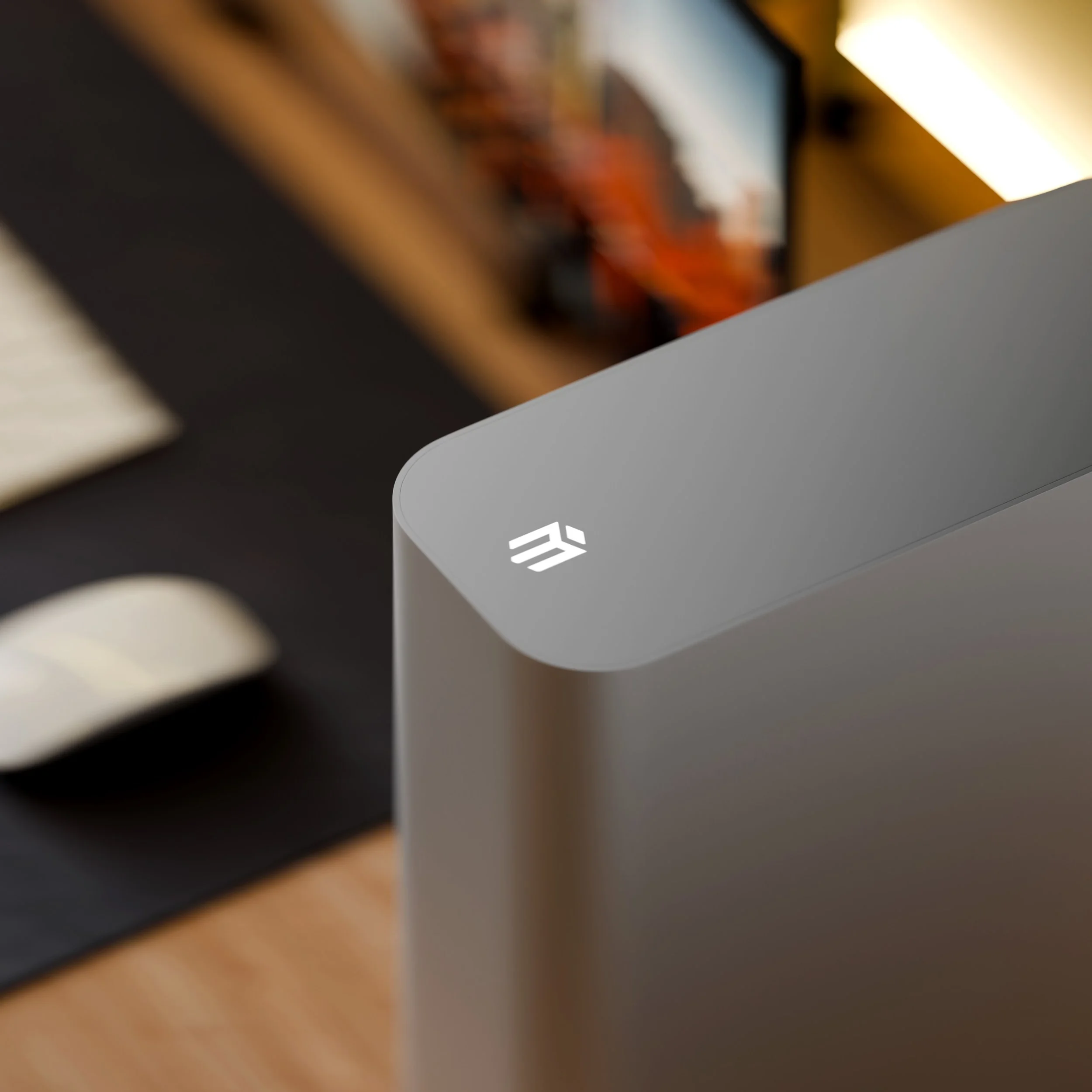 Close-up of a silver electronic device with a logo on top, placed on a wooden desk. In the background, there is a blurred white mouse, a keyboard, and a shelf with books.