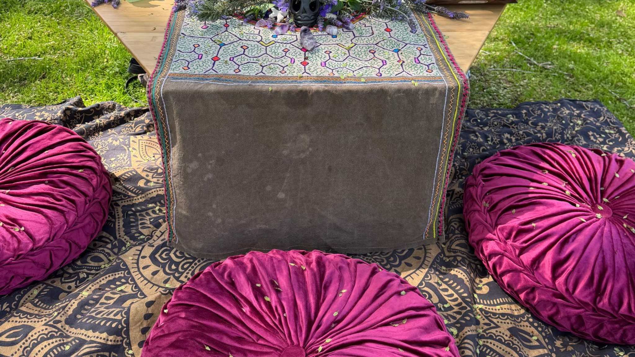 Outdoor setting with a table covered in decorative cloth, purple round cushions, and a patterned rug on grass.