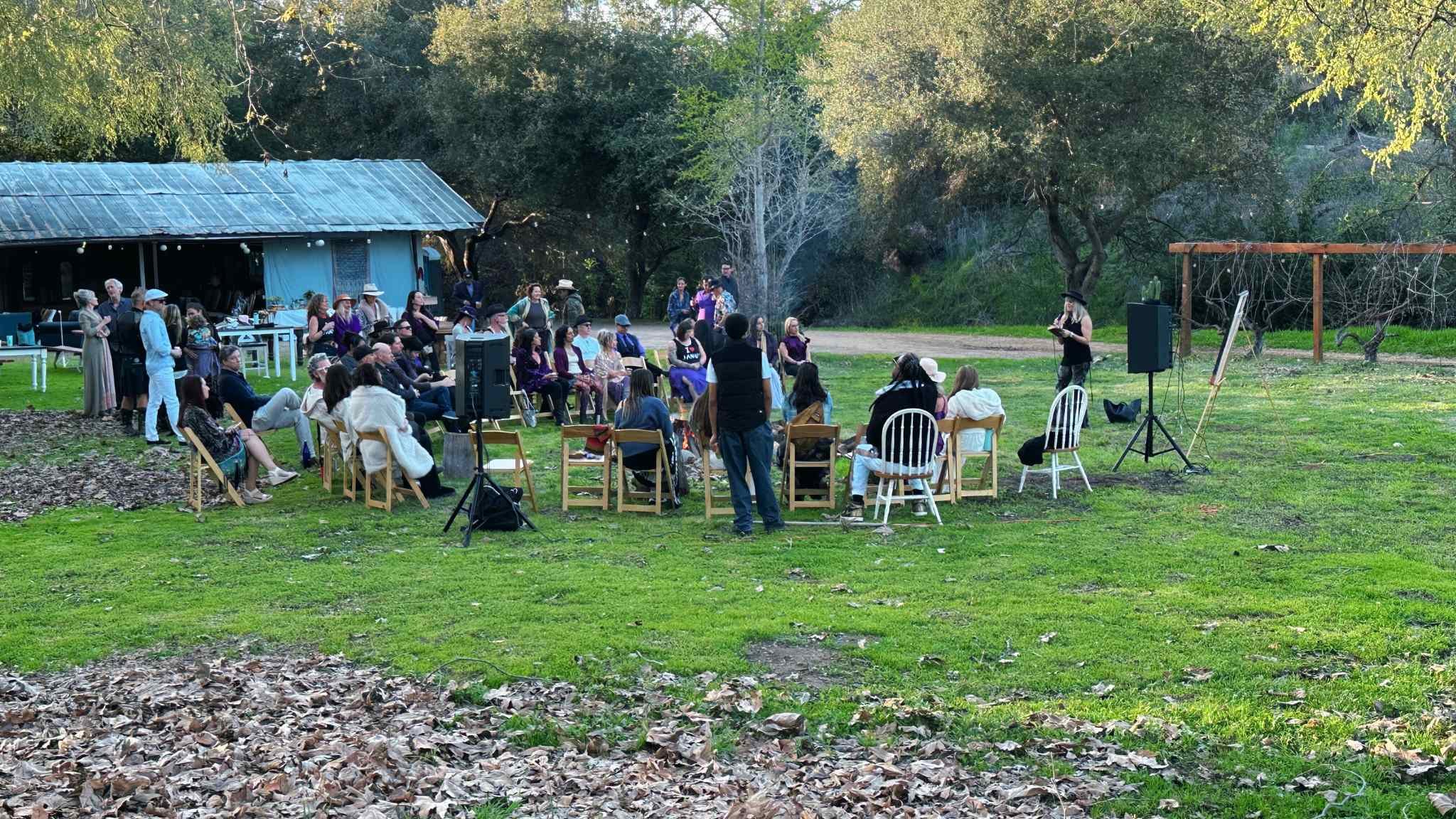 Outdoor gathering with people seated on chairs in a circle, listening to a speaker in a natural setting with trees and a blue building in the background.