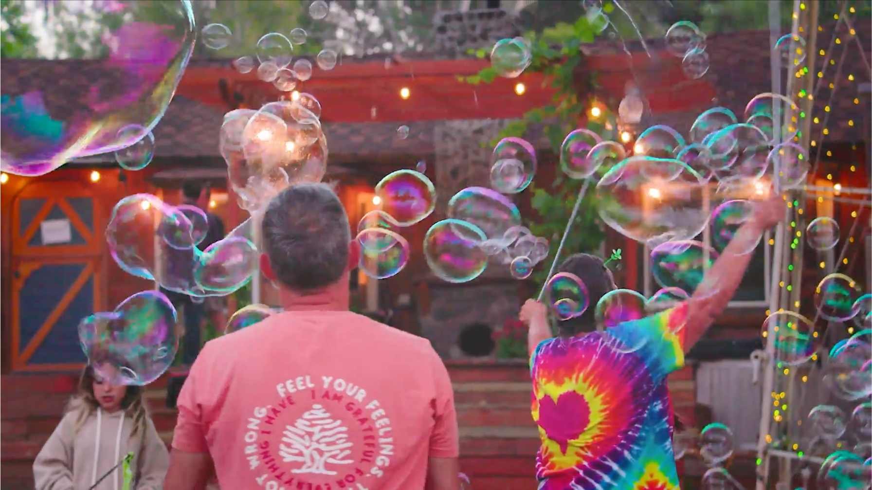 People creating and playing with large soap bubbles, outdoors, wearing colorful shirts and surrounded by lights.