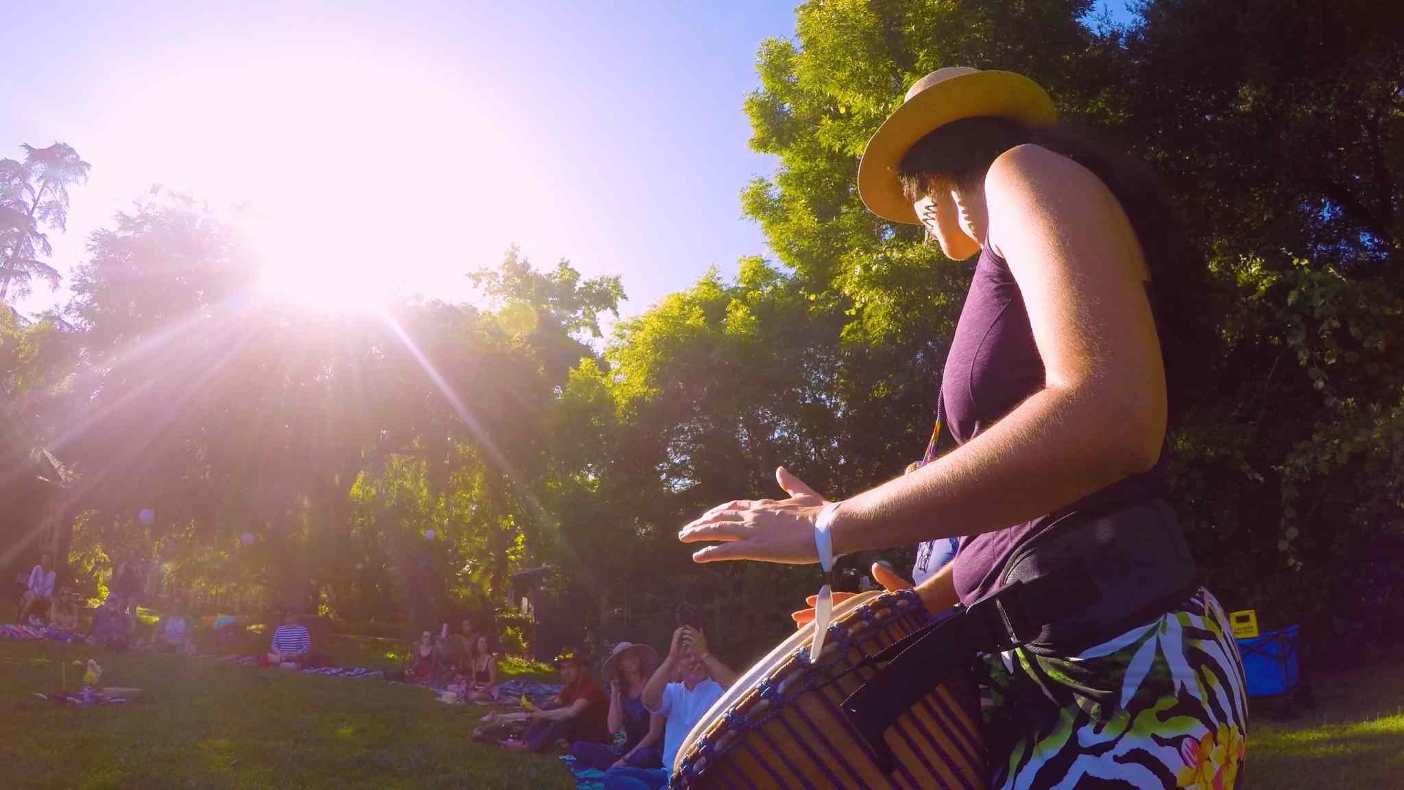 Person playing a drum outdoors with sunlight and trees in the background