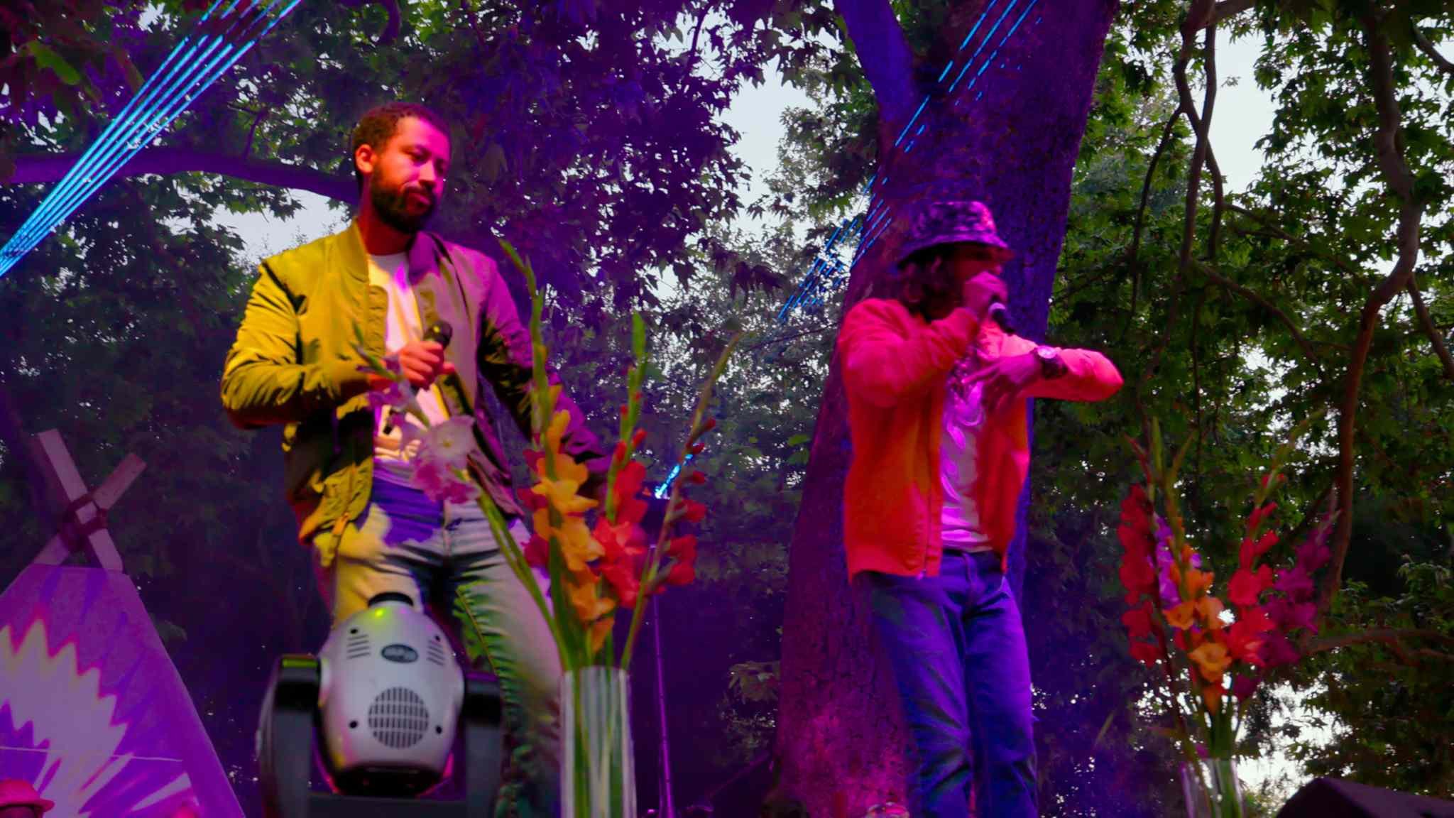 Two men performing on an outdoor stage with colorful lighting and trees in the background. One is wearing a green jacket and the other a red one, both holding microphones. Vibrant flowers are in the foreground.