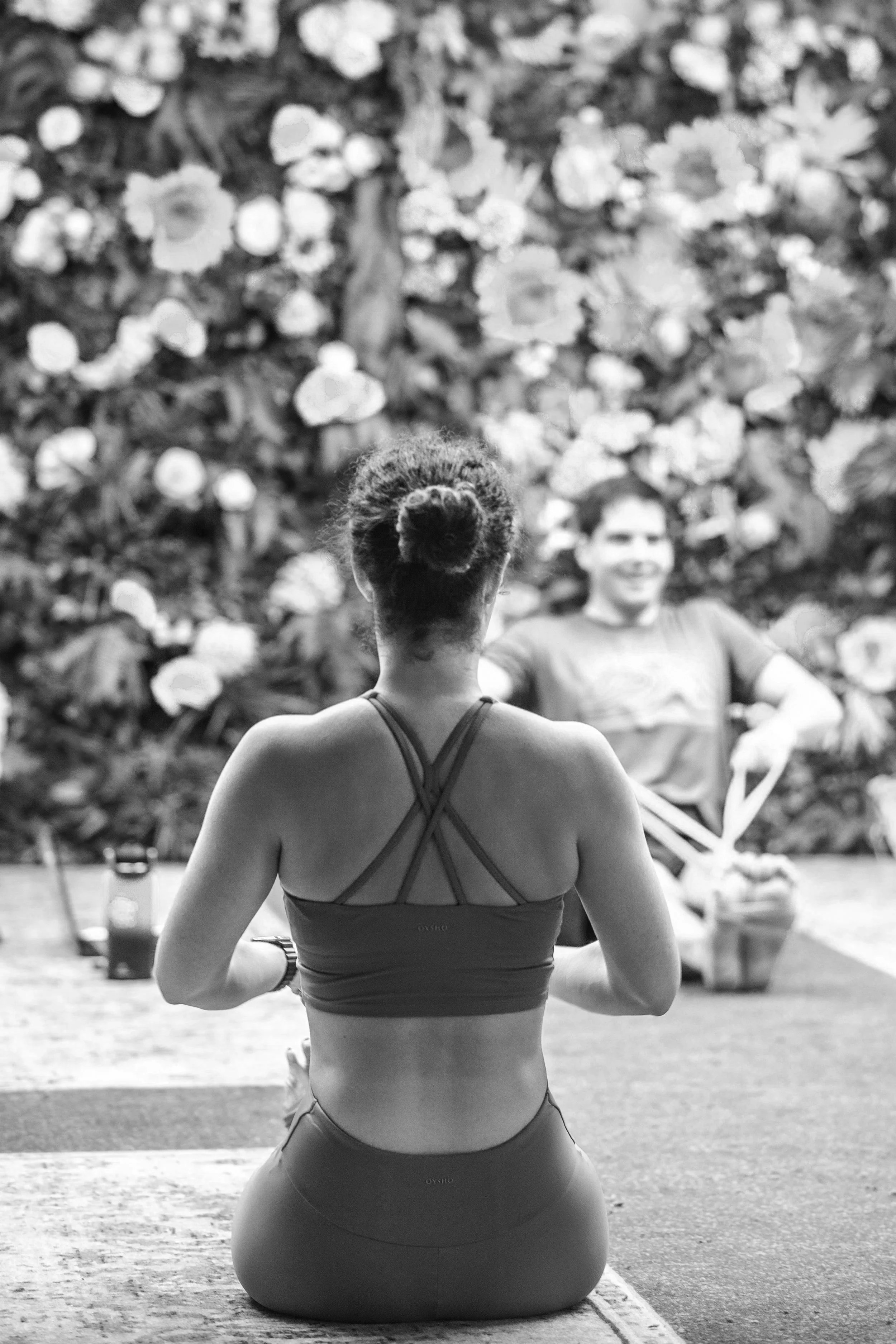 A woman practicing yoga or meditation outdoors, seen from behind, with a man sitting cross-legged in front of a floral backdrop.