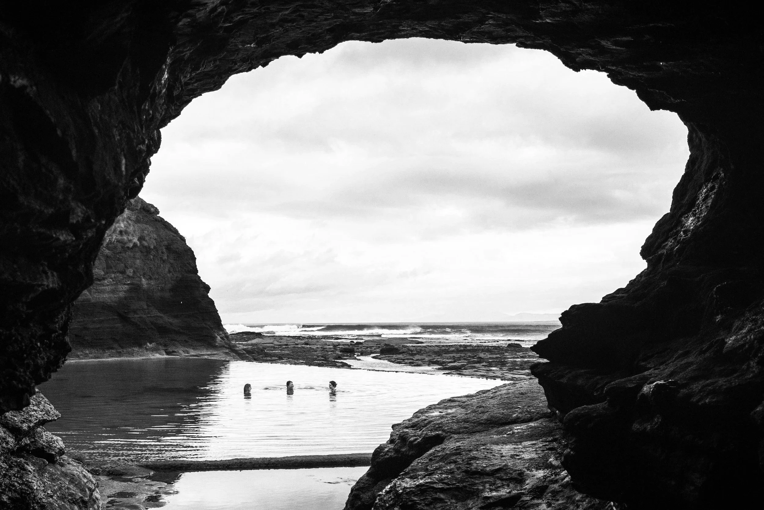 A black-and-white photograph of a rocky coastal scene viewed through a natural cave opening. The image shows a shoreline with a small body of water, distant waves, and three people wading in the water near the beach.
