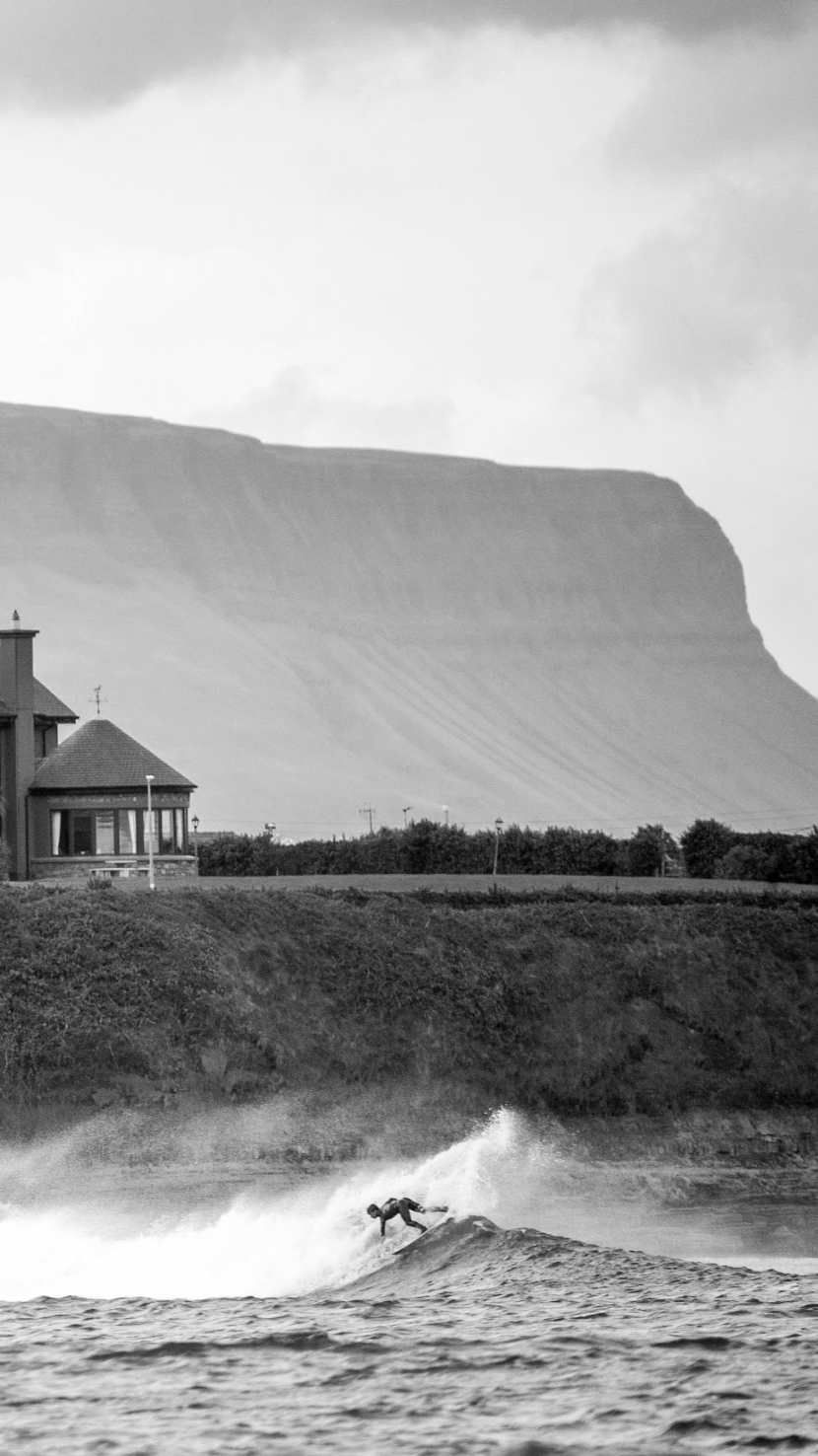 A person surfing on a wave near a shoreline with a house and a mountain in the background, black and white photo.