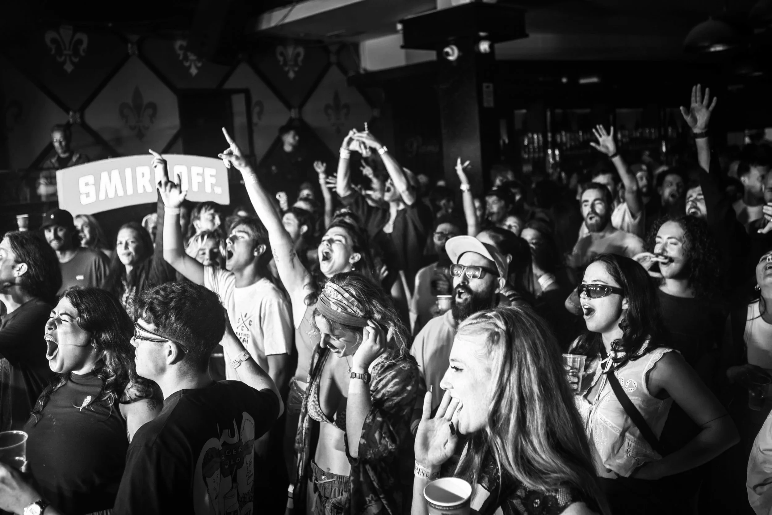A crowd of people at a concert or music event, cheering and singing passionately, with some wearing sunglasses and holding drinks. There is a sign with the text 'SMIRNOFF' in the background.