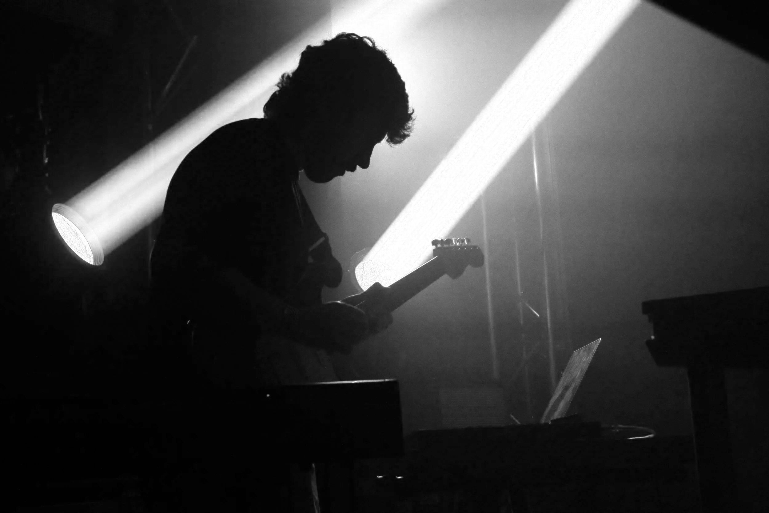 Silhouette of a person playing an electric guitar on stage with a bright light shining behind them in a dark setting.