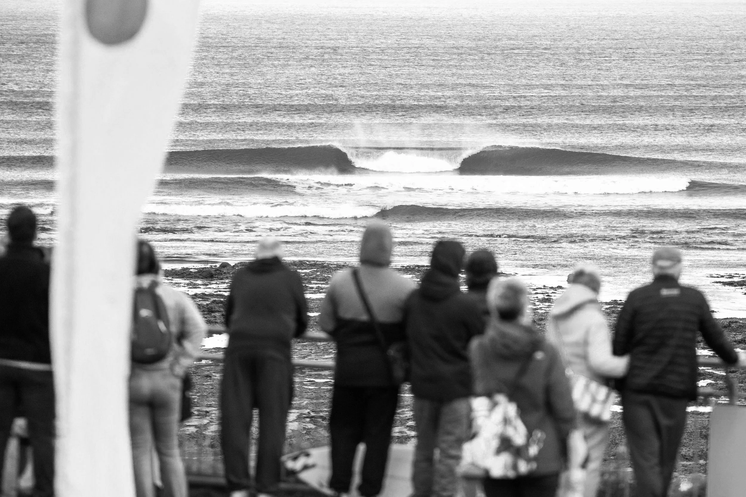 A group of people standing on a rocky beach, watching large ocean waves and a surf break in the distance.