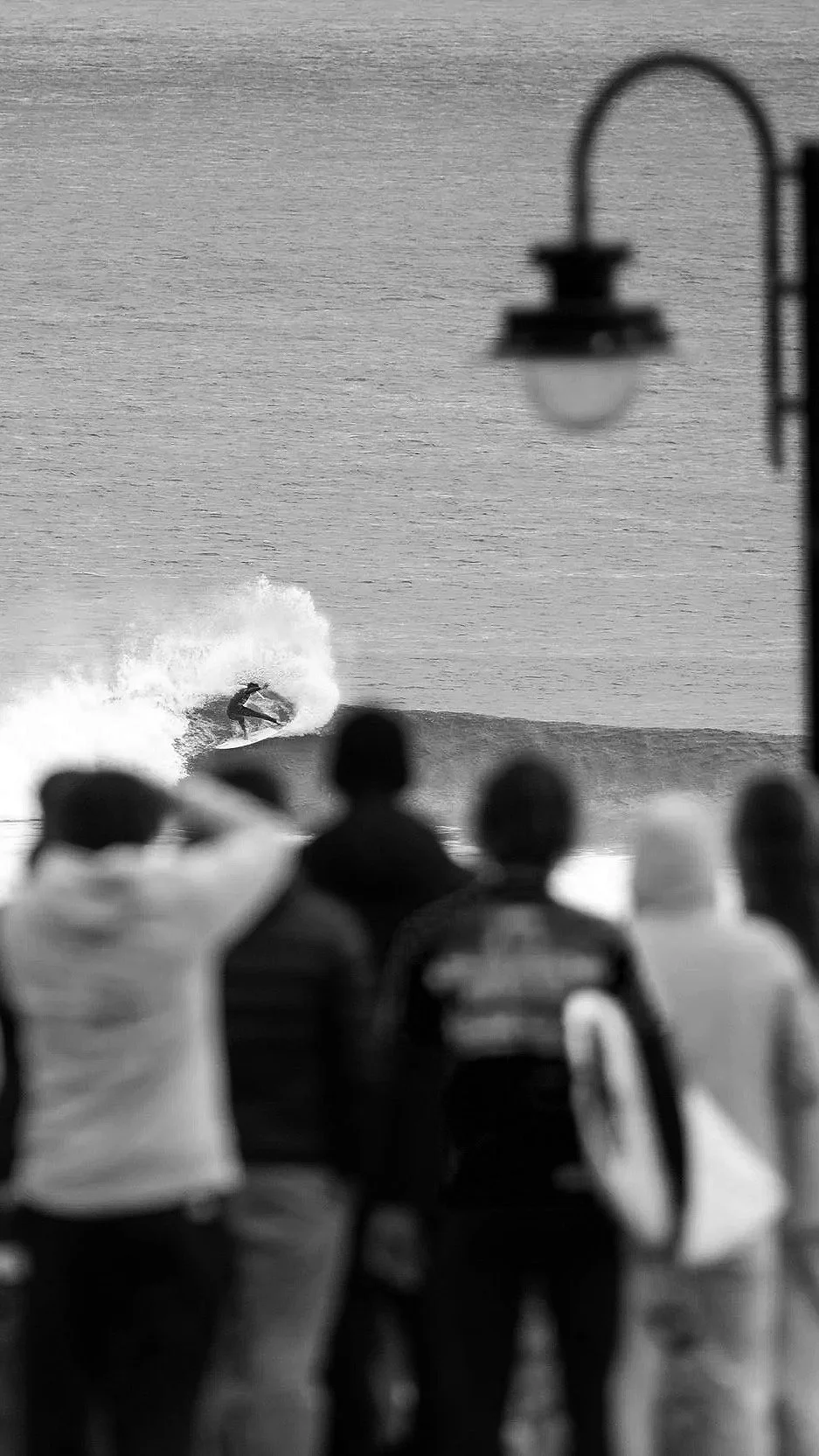 A surfer riding a wave at sunset seen from a balcony with people watching, and an overhanging street lamp in the foreground.