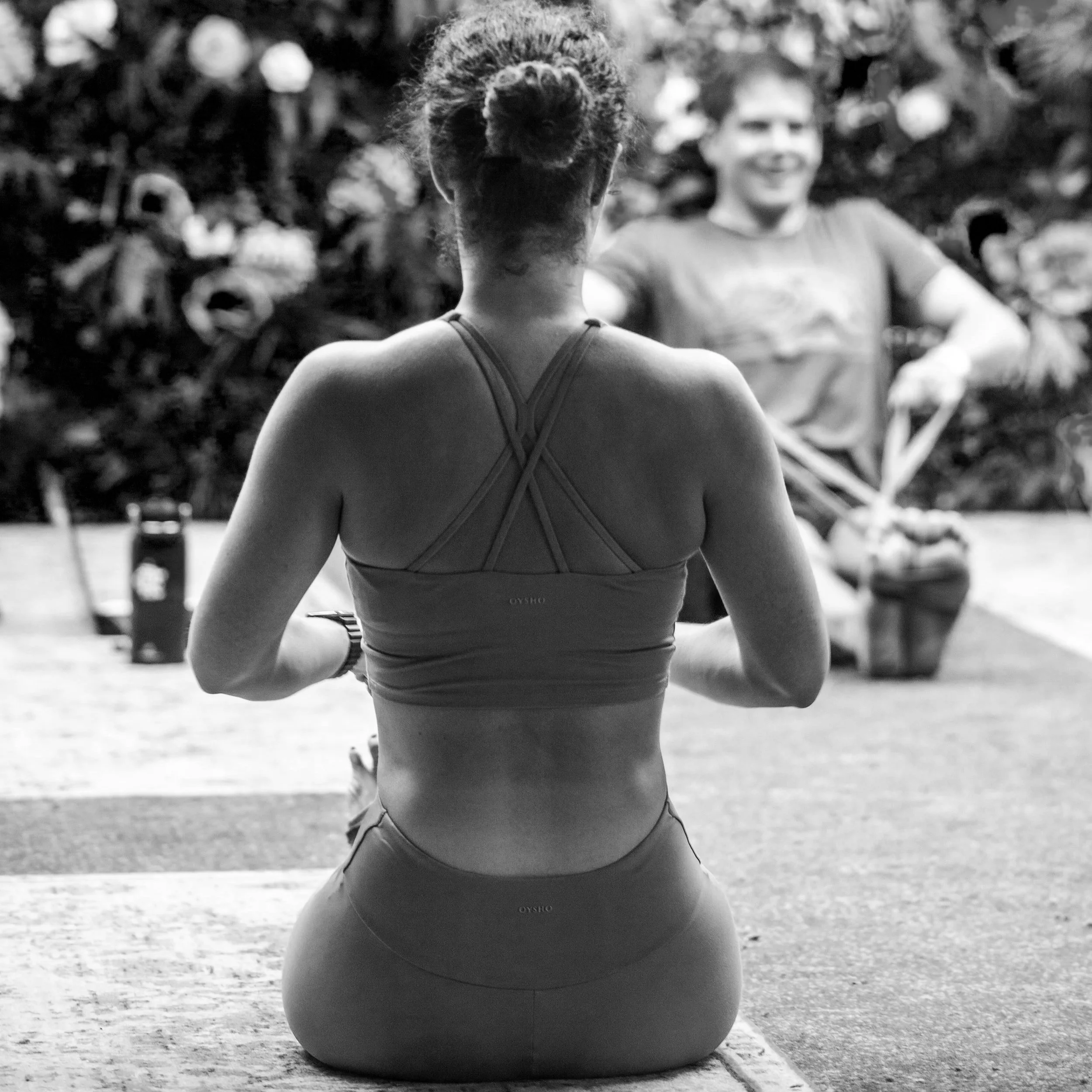 Backside of a woman with braided hair sitting on a yoga mat, wearing a sports bra and leggings, in a workout or yoga studio with a man in the background holding a plant and smiling.