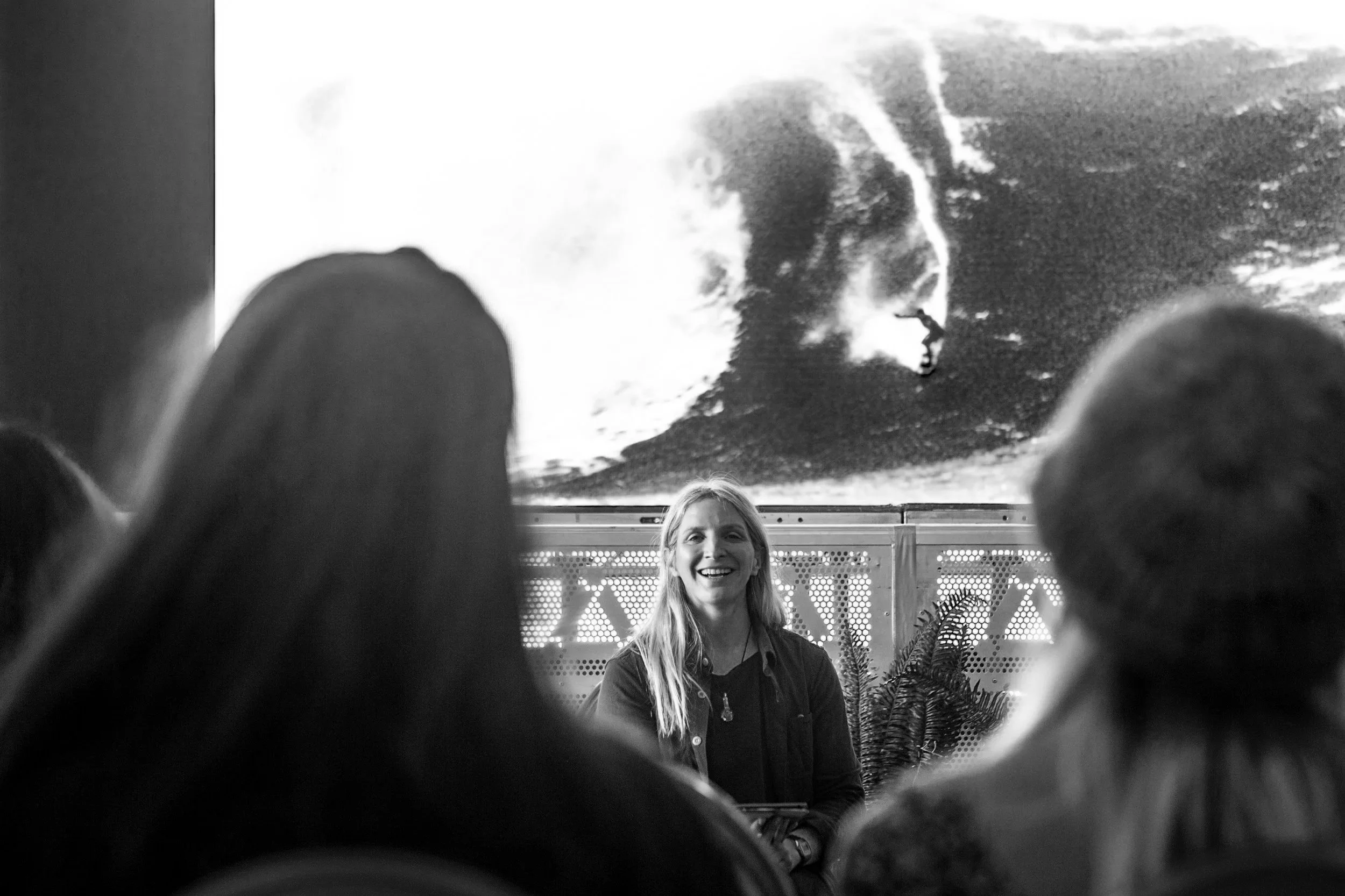 A woman smiling while presenting in front of a large screen showing a black and white image of a huge wave.