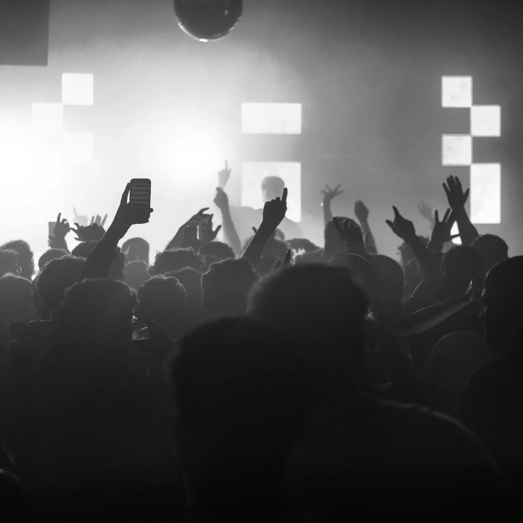 Black and white photo of a crowded concert or club with people dancing and raising their hands, some holding phones, with a bright light and geometric screens in the background.