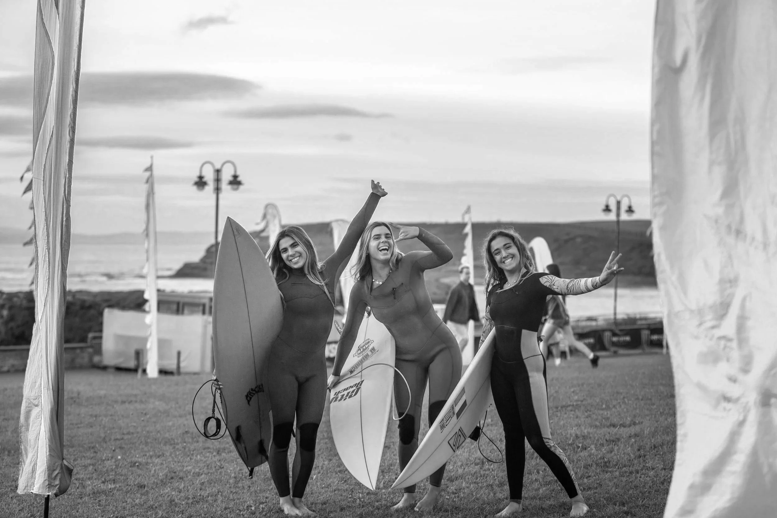 Three women in wetsuits holding surfboards on a beach with flags and lamps in the background, smiling and celebrating.