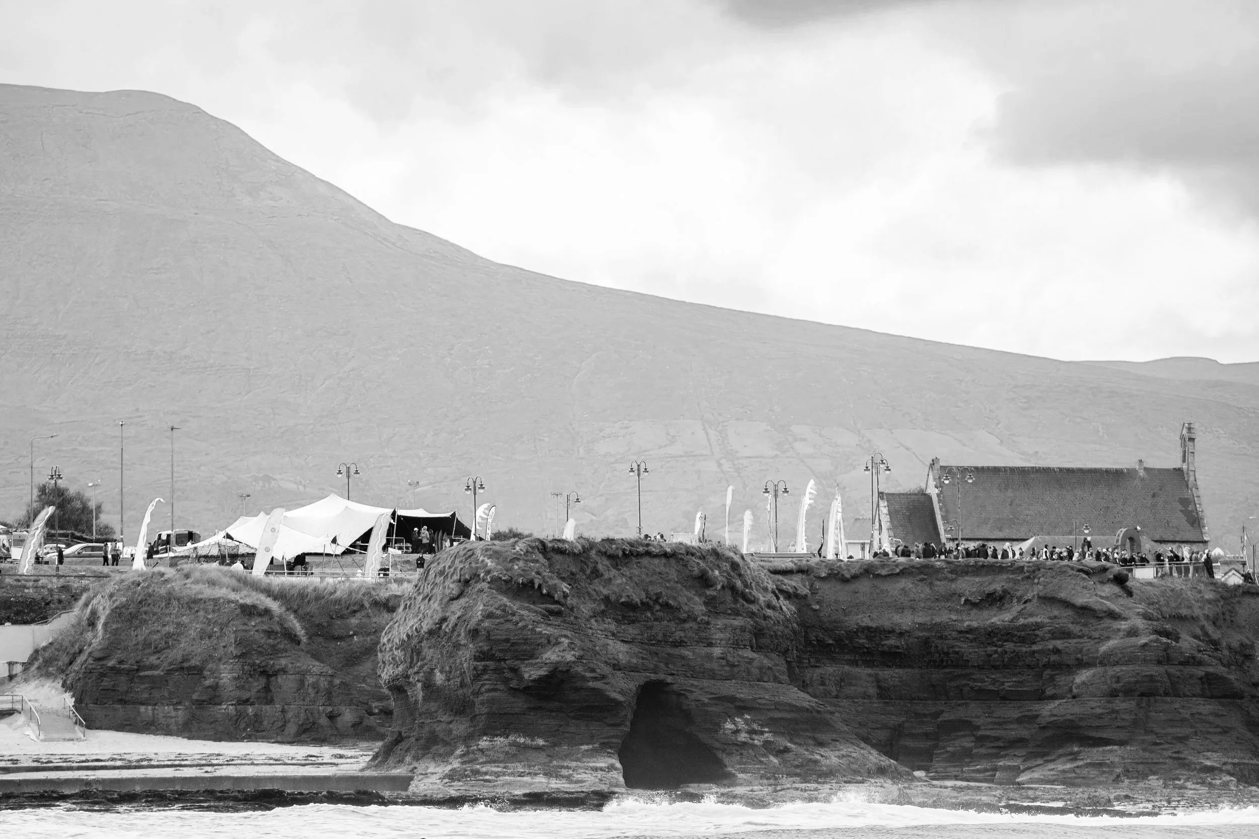 Black and white photo of a coastal area with rock formations in the foreground, tents and a small building on the shoreline, and hills in the background.