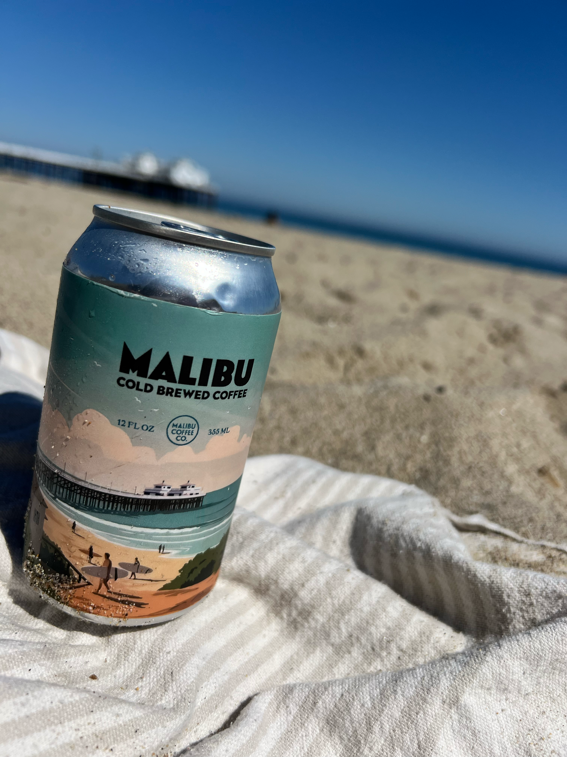 A can of Malibu cold brewed coffee is resting on a white cloth at the beach with sand and the ocean in the background.