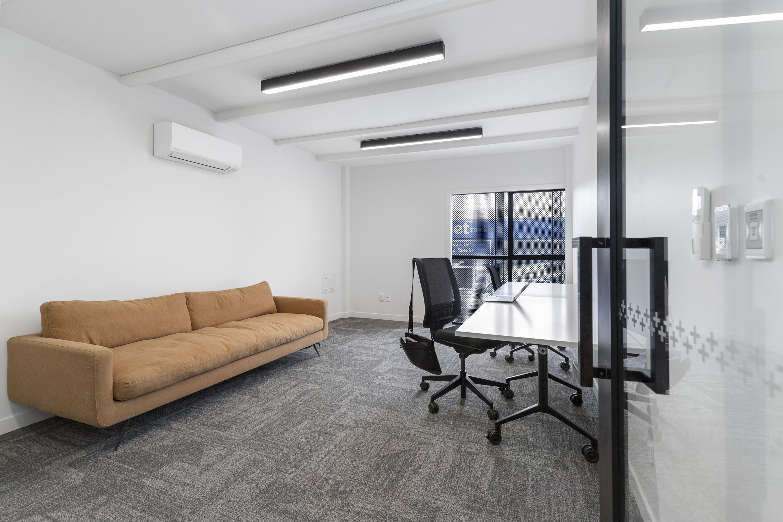 Modern office with a brown sofa, two black office chairs, a white table with a laptop, carpet flooring, and a glass wall.