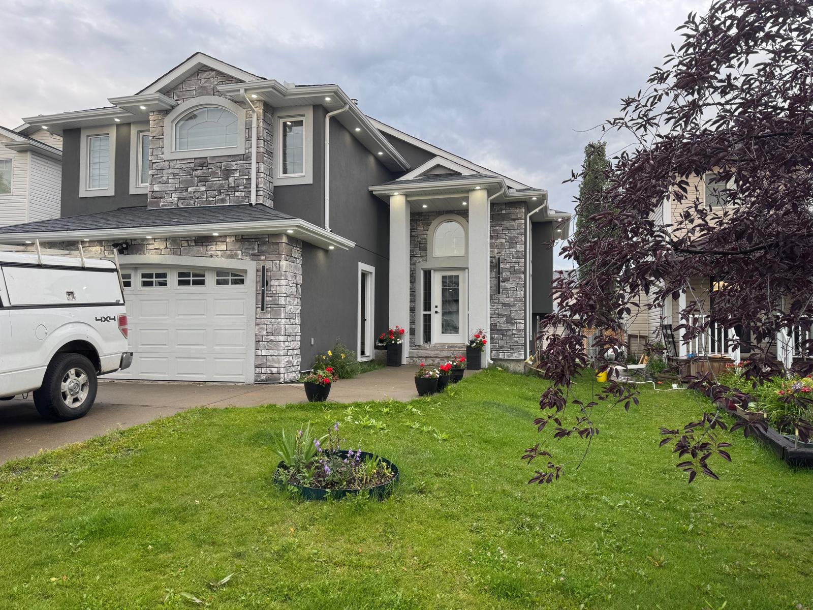 A modern two-story house with a stone and gray exterior, white trim, and a garage door. The front yard has a green lawn, potted flowers, and a driveway with a white pickup truck parked.