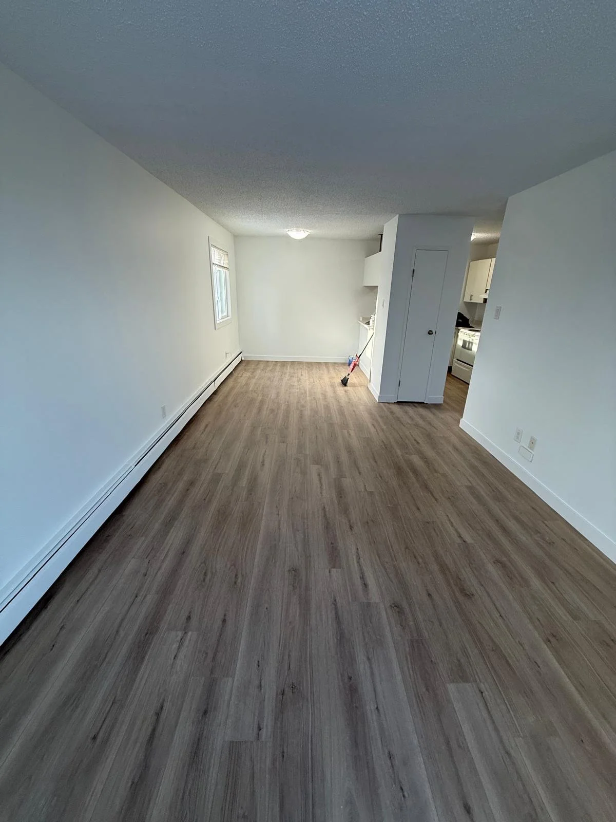 Empty living room with wood laminate flooring, white walls, a window, and a small kitchen area visible in the background, with a broom leaning against the wall.
