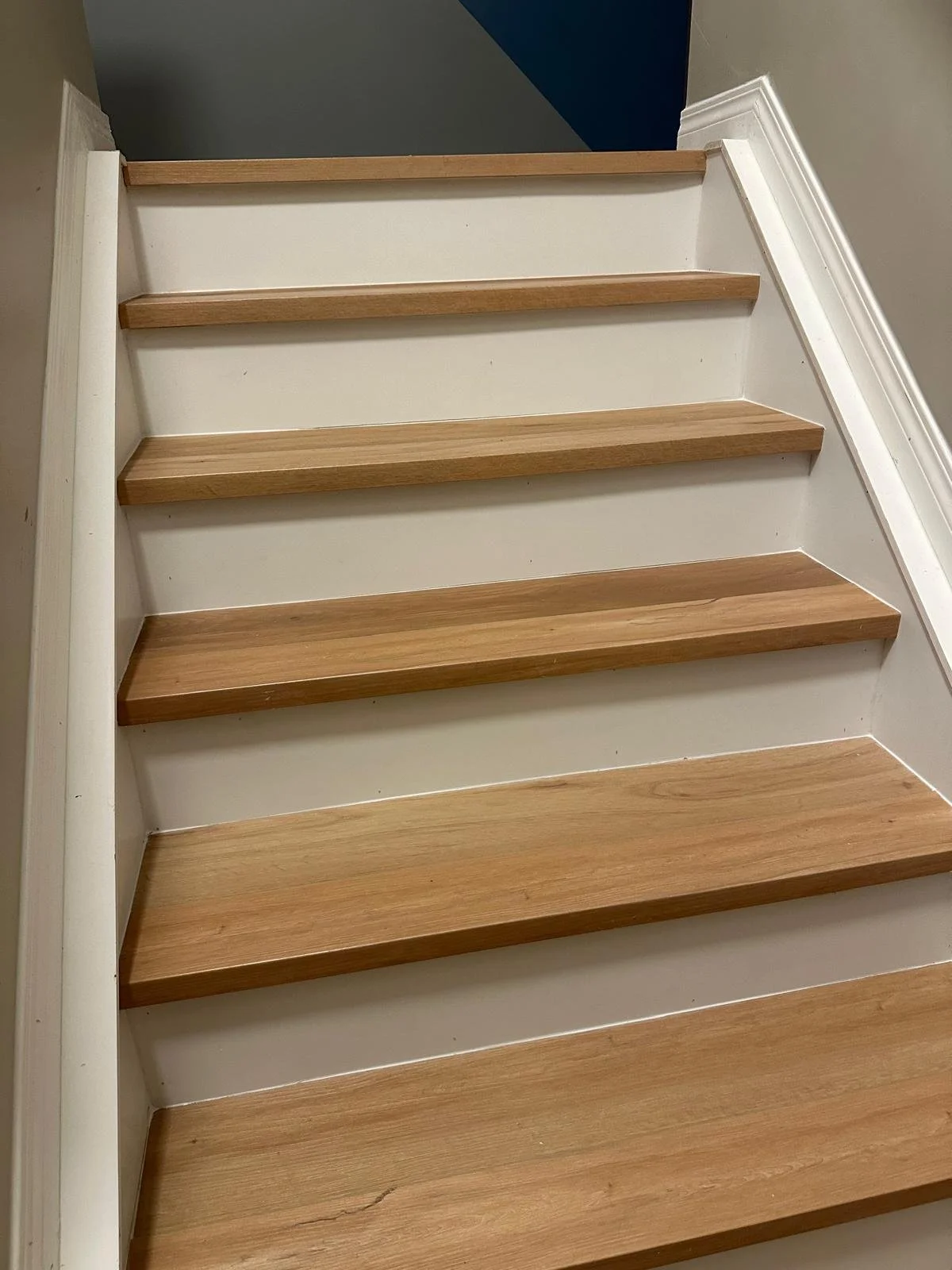 A set of wooden stairs with beige risers and light brown treads, next to a white wall with a decorative white baseboard.