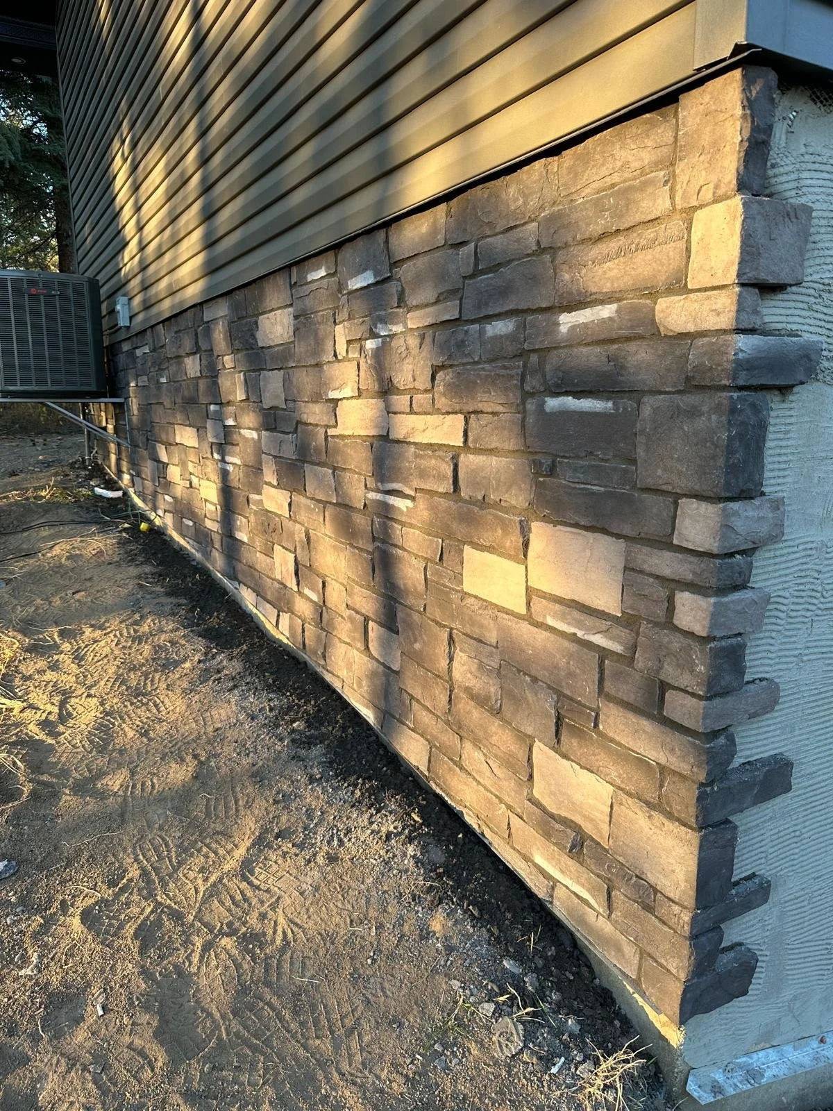 Exterior wall of a building with stonework on the lower half and horizontal siding on the upper half, with an air conditioning unit on the left side.