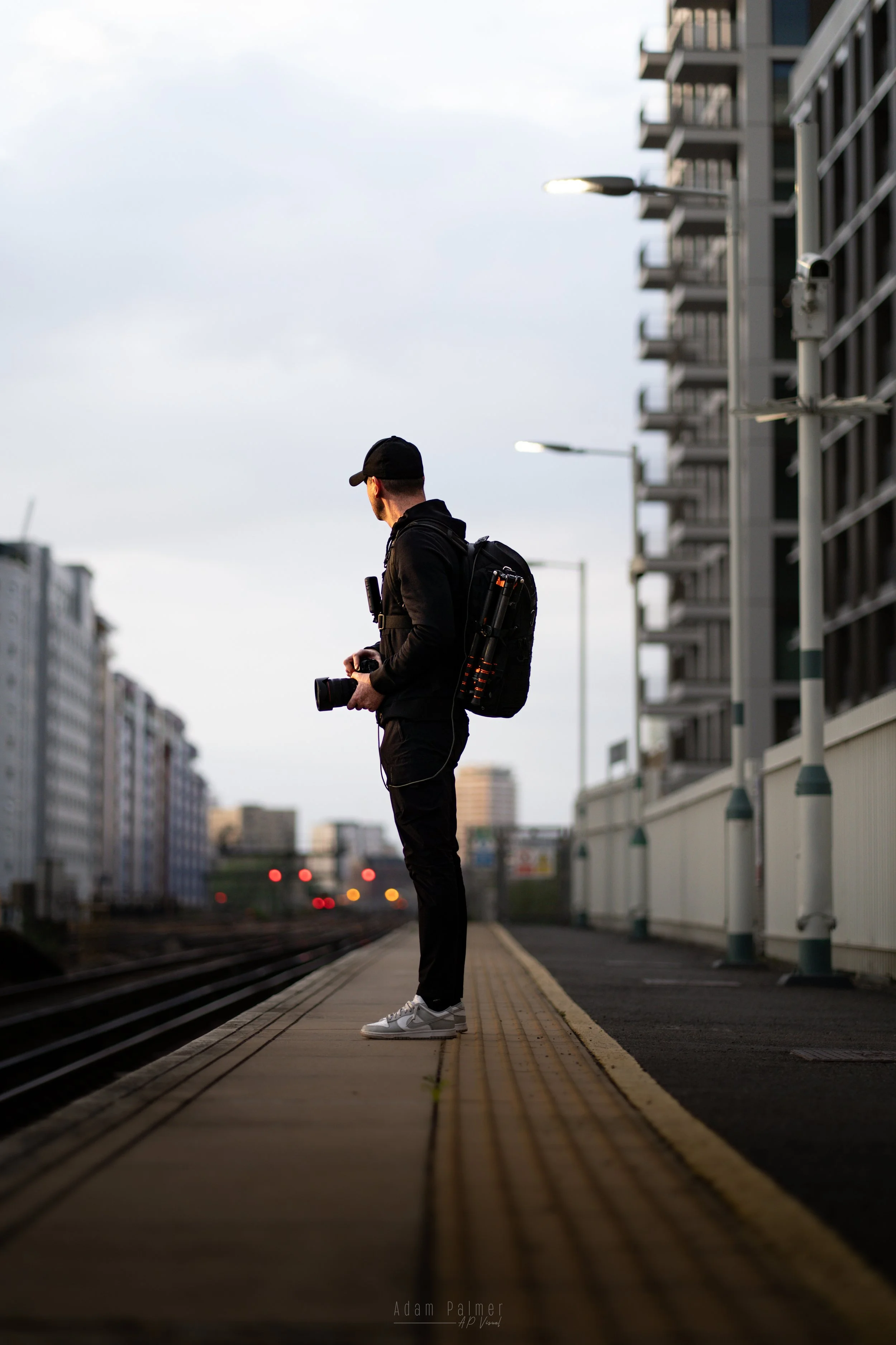 A man standing on a train platform holding a camera with start of cityscape in background.
