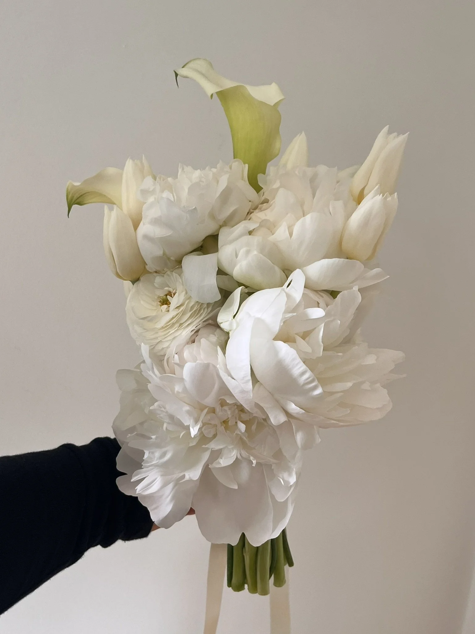 A bouquet of white flowers, including calla lilies, peonies, and tulips, held by a person wearing a black sleeve against a plain beige wall.