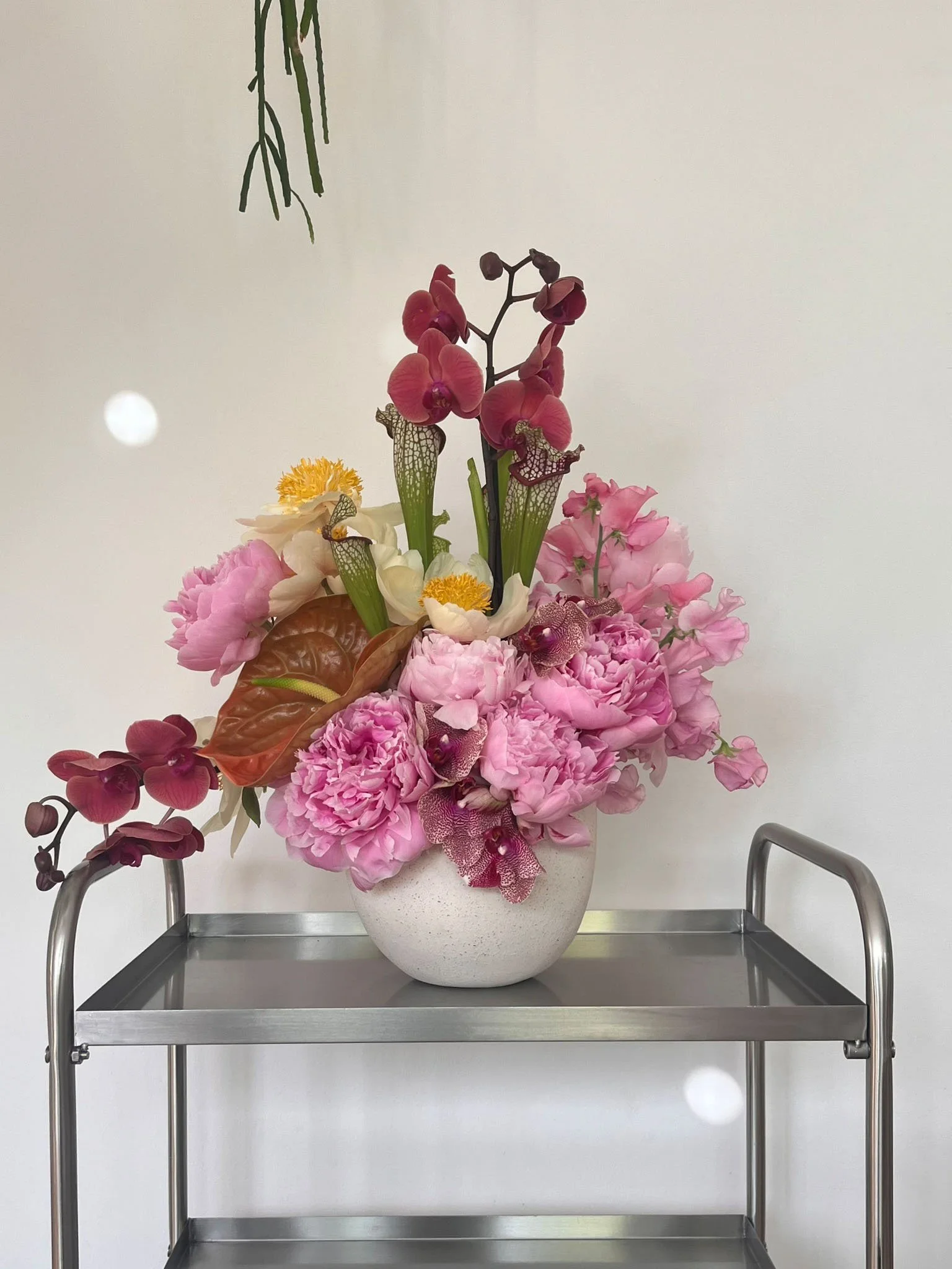 A large flower arrangement with pink peonies, orchids, and other blossoms in a white vase on a stainless steel cart