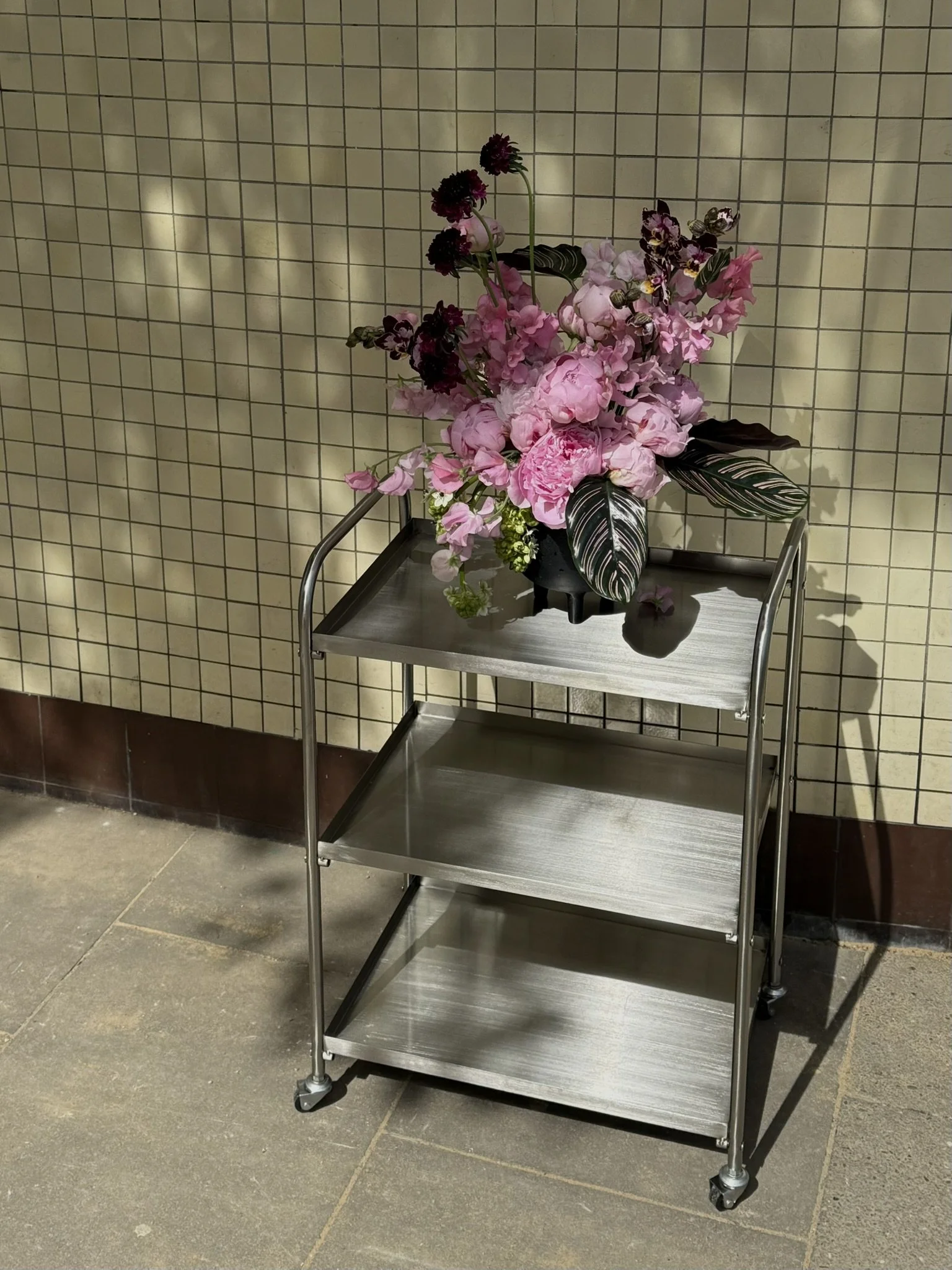 A metal cart with three shelves holding a flower arrangement with pink peonies, purple and black flowers, and variegated green and white leaves, against a beige tiled wall.
