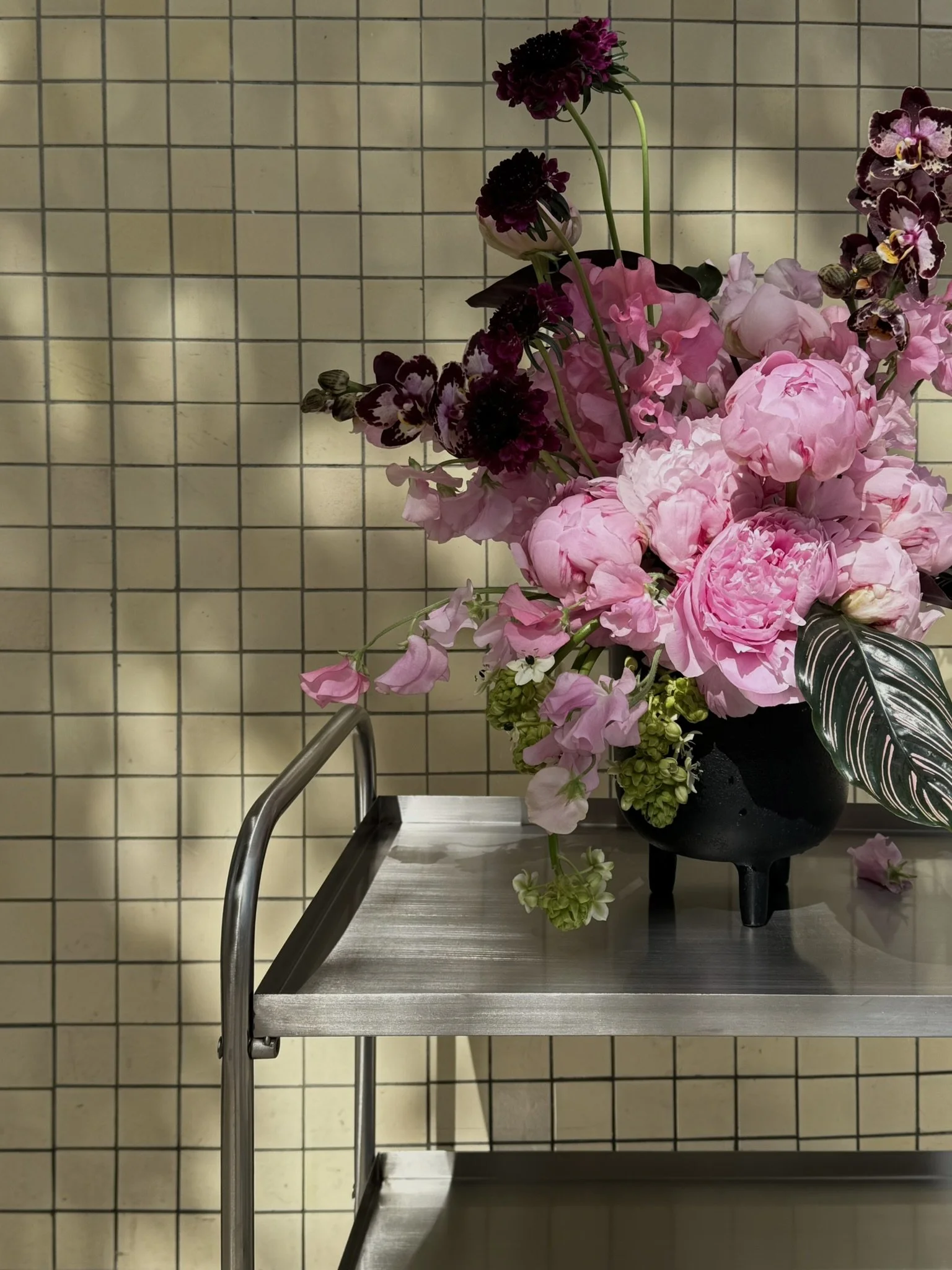 Vase with pink and purple flowers on a stainless steel table against a beige tiled wall.