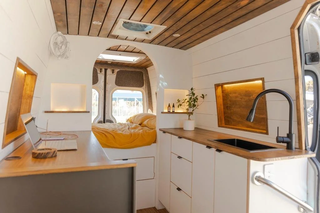 Interior of a tiny camper van with white walls and wooden accents, a small kitchen with a black sink and a faucet, and a bed area with a yellow blanket, visible through an archway.