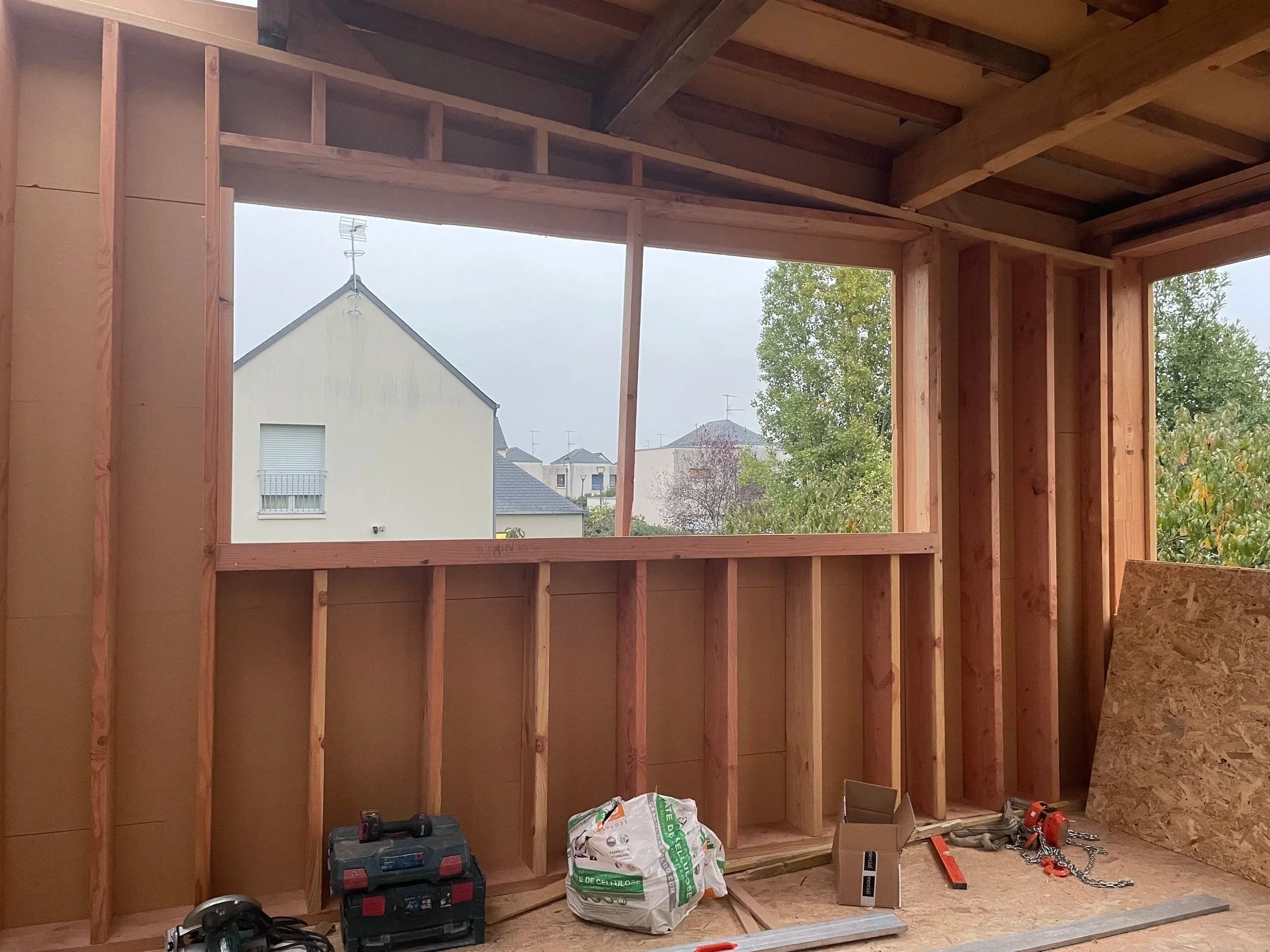 Intérieur en construction avec structure en bois, fenêtre ouverte à l'extérieur, outils et matériaux de construction sur le sol.