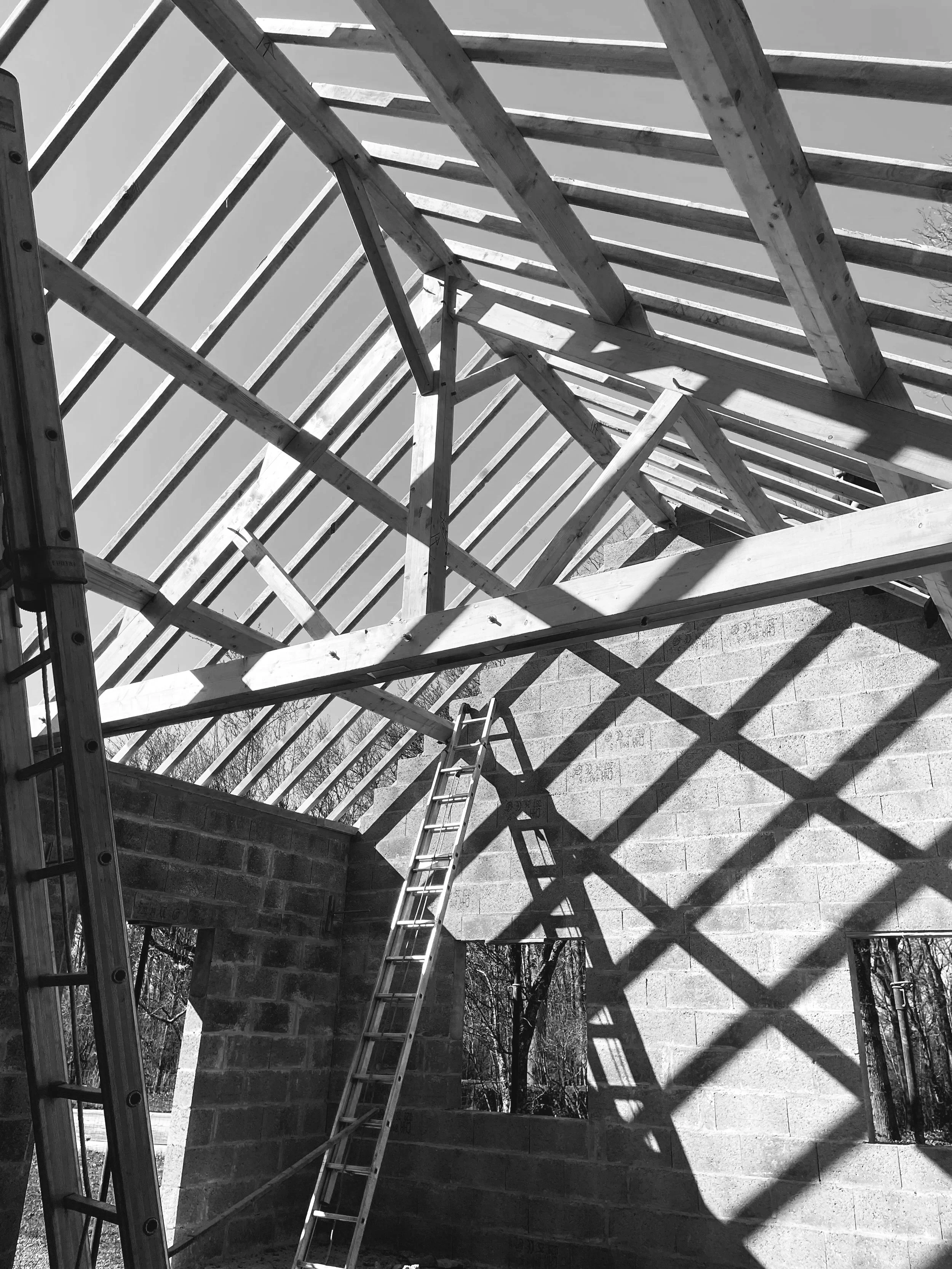 Structure en bois en construction, avec des poutres et des chevrons, et de longues ombres projetées par le soleil sur un mur en parpaings.