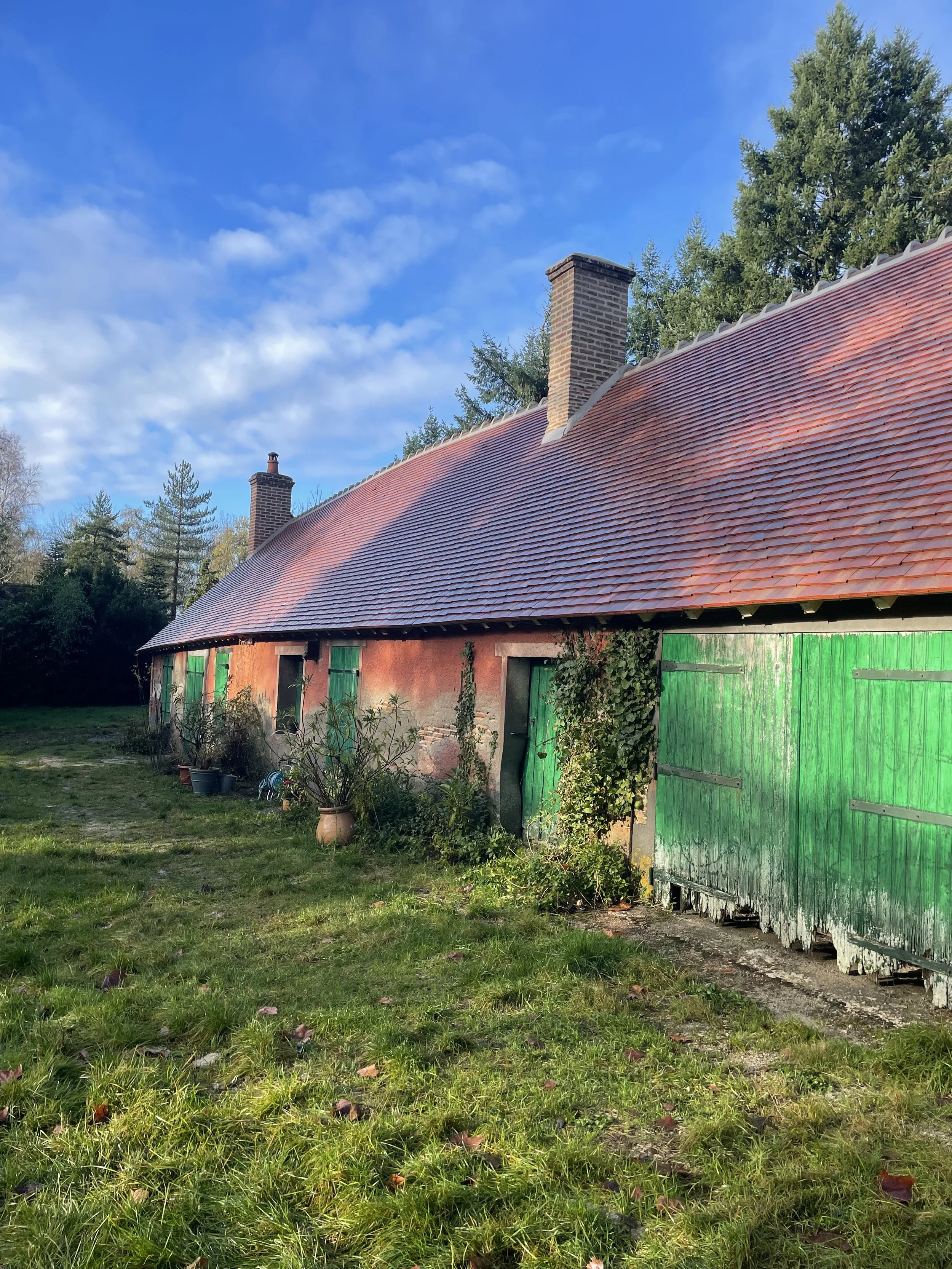 Une vieille maison en pierre avec des volets verts, un toit en tuiles rouges, entourée d'un jardin avec des pots de fleurs et des arbres en arrière-plan, sous un ciel bleu avec quelques nuages.