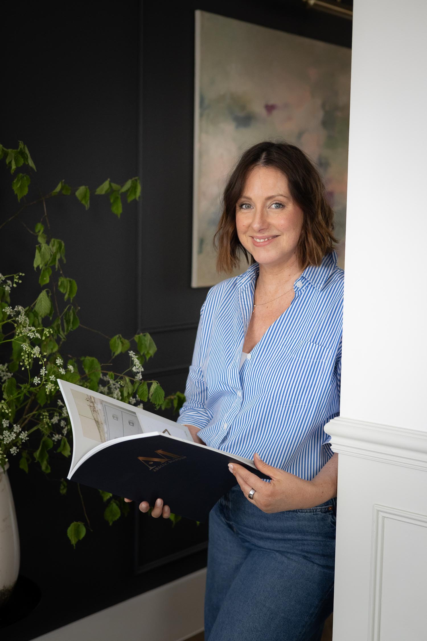 Woman with shoulder-length brown hair wearing a blue and white striped shirt, holding an open magazine, and smiling while standing near a white wall with artwork in the background and a plant nearby.