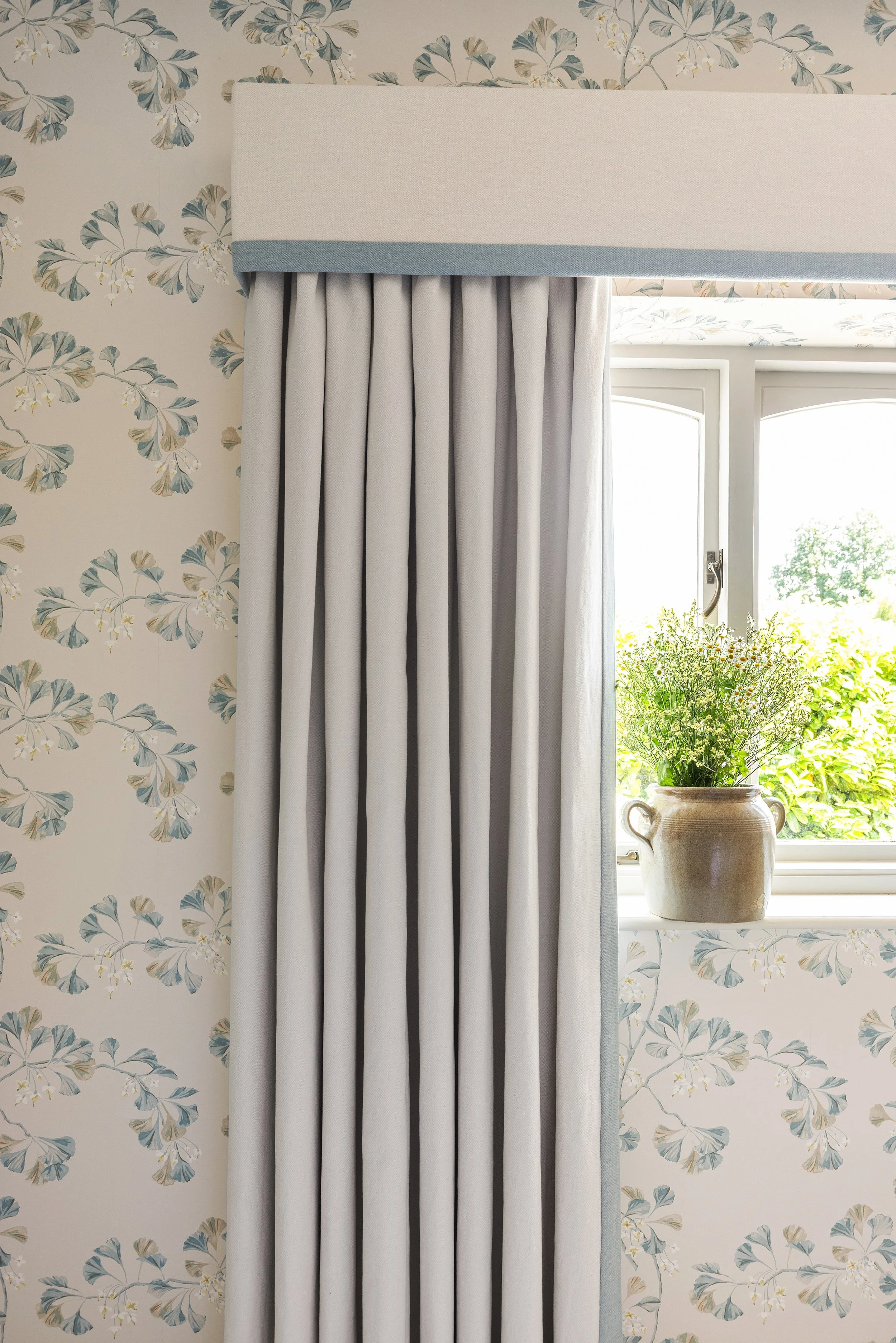 A window with bespoke handmade curtains, and a plant in a rustic pot on the windowsill, with floral wallpaper in the background.