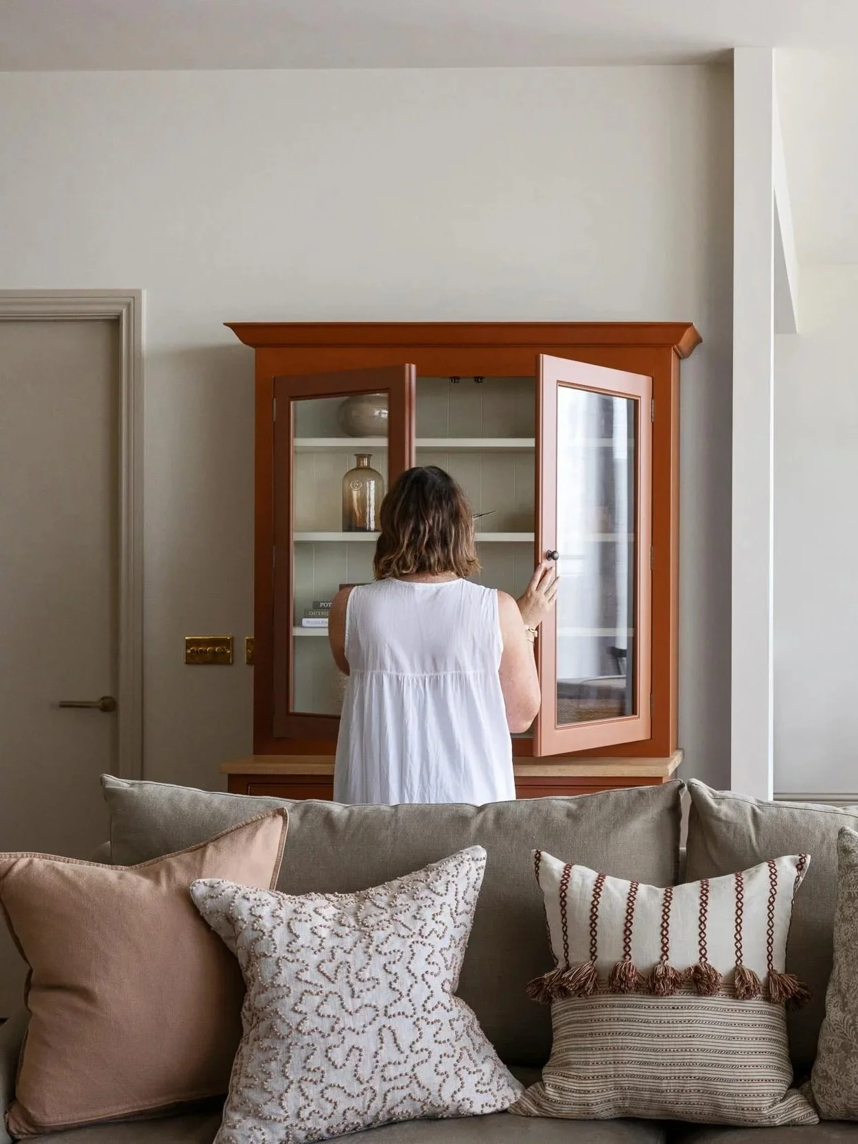 A woman in a white sleeveless dress opens a wooden display cabinet in a living room with a beige sofa and decorative pillows in front of her.