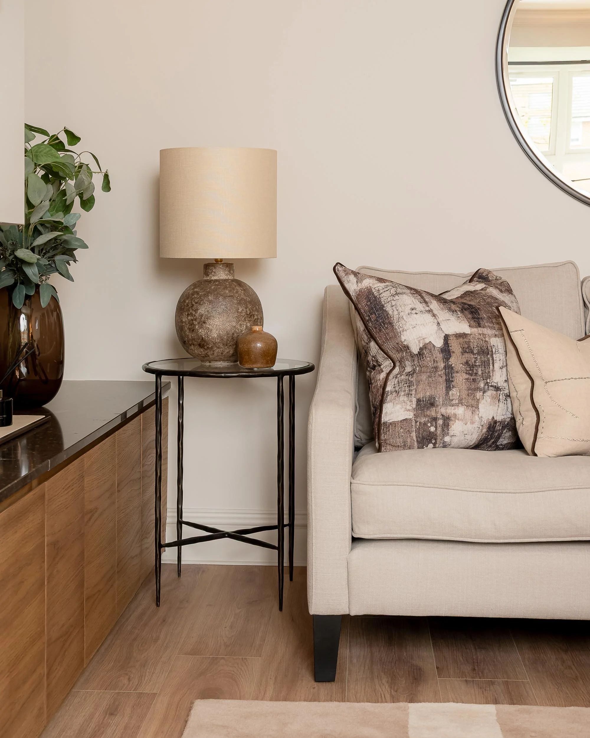 Side table with two decorative pots and a beige lampshade next to a beige sofa with patterned and plain pillows, against a white wall with a round mirror.