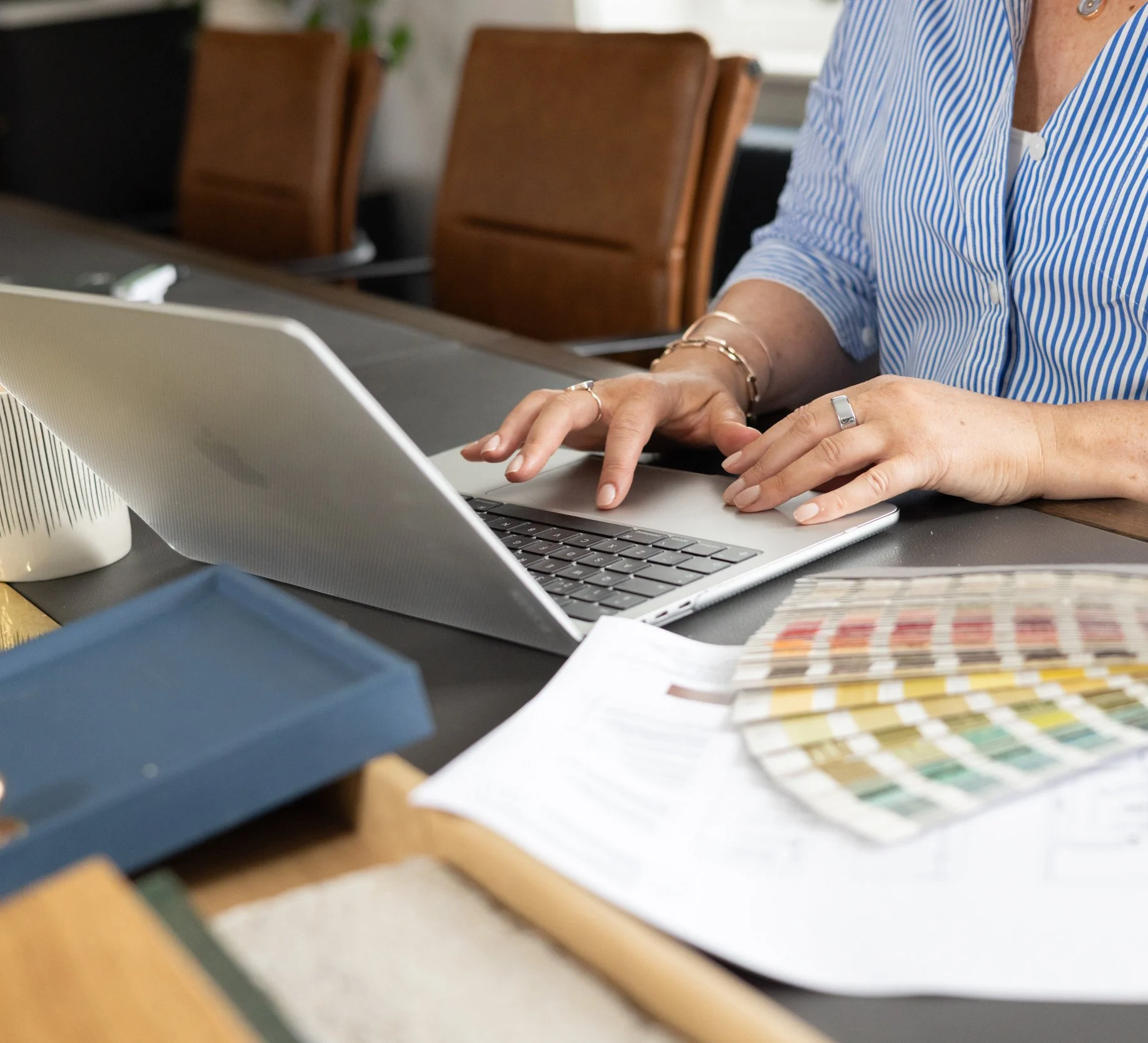 A woman working on a silver laptop at a desk, with color swatches and design samples nearby.