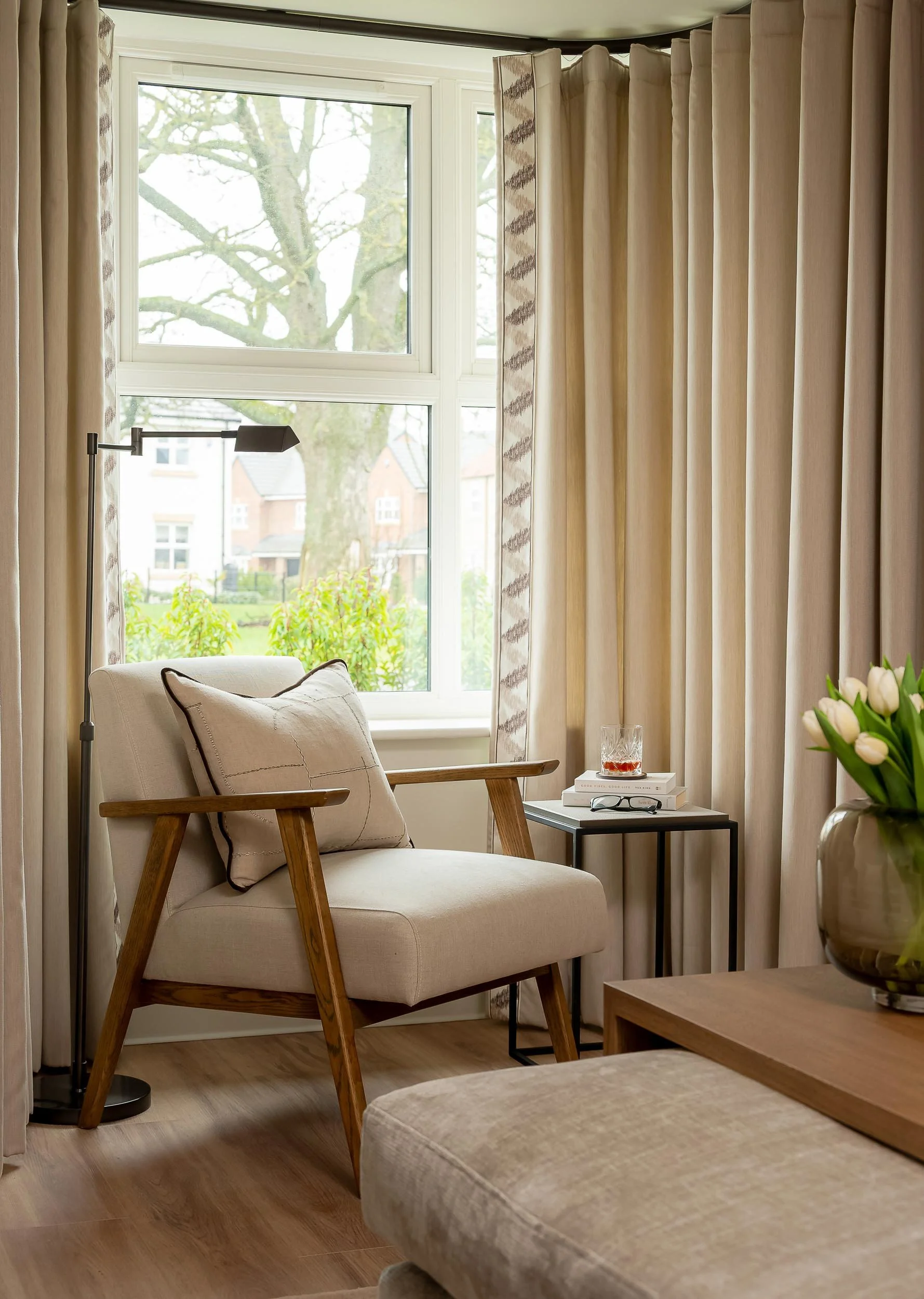 A cozy living room corner with a beige armchair, a small black side table with books and a glass of drink, a vase with white tulips, large window with beige curtains, and a view of trees and houses outside.