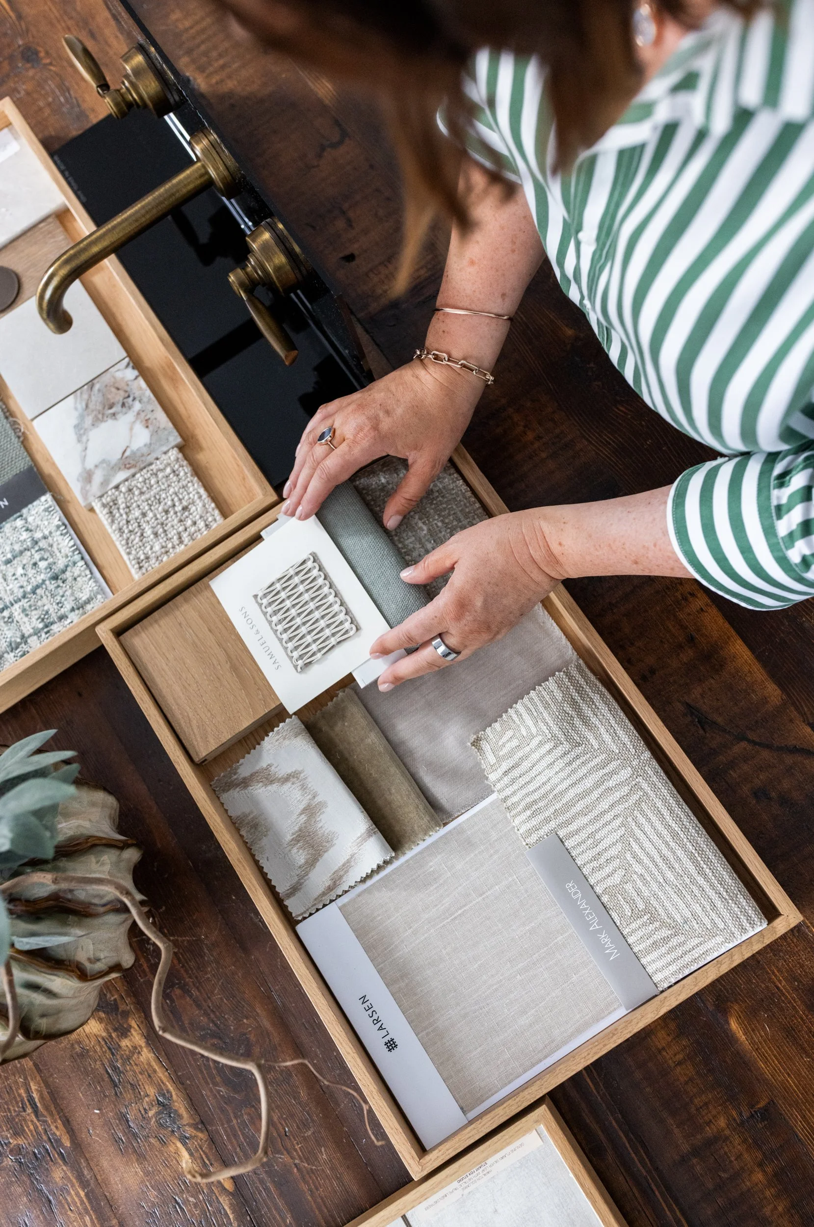 Person in a green and white striped shirt organizing fabric samples in a wooden drawer, with other swatches and fabric samples nearby.