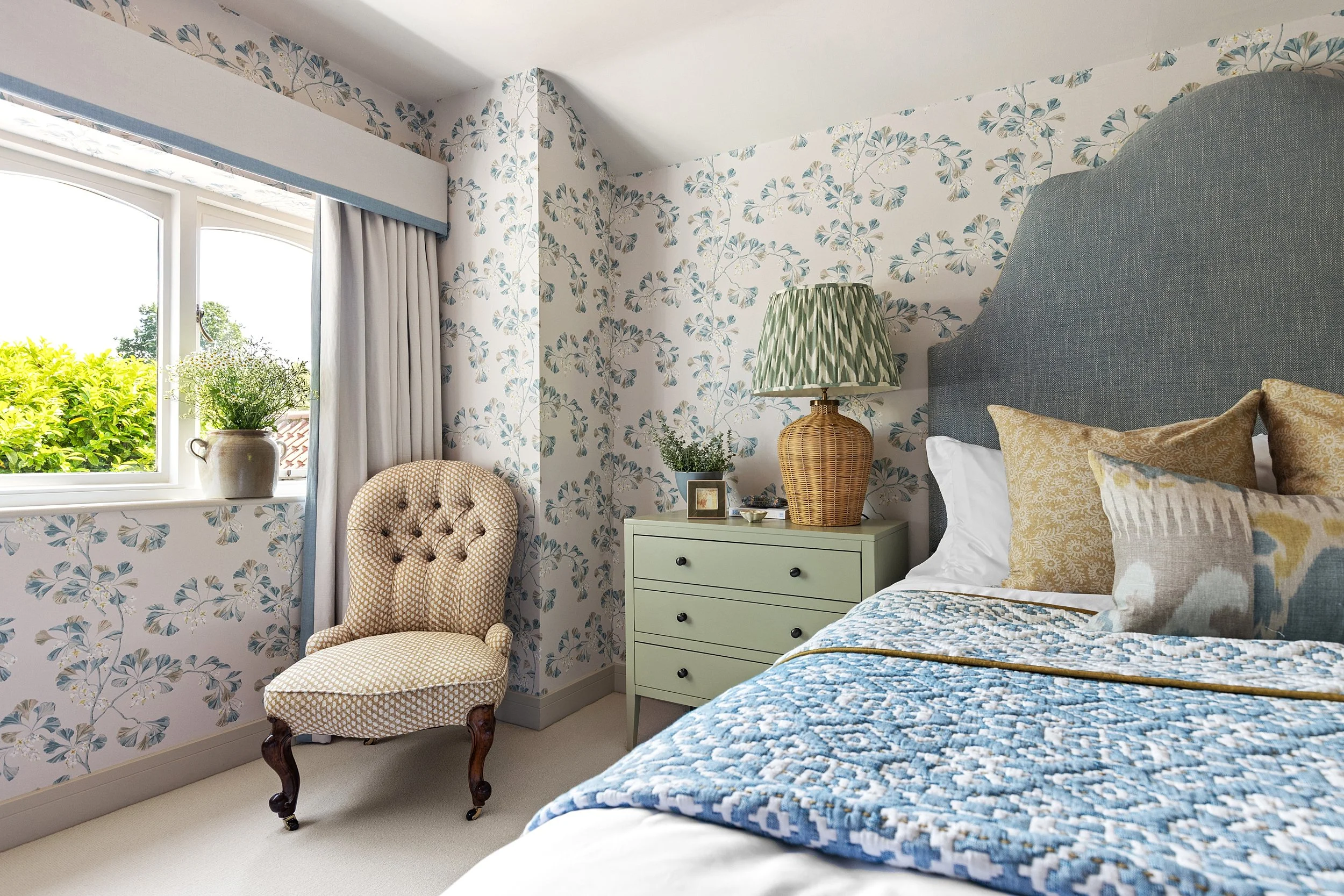 A cozy bedroom with floral wallpaper, a window with a plant on the sill, a beige upholstered armchair, a light green dresser with a wicker lamp and small plants, and a bed with patterned pillows and a quilted blanket.