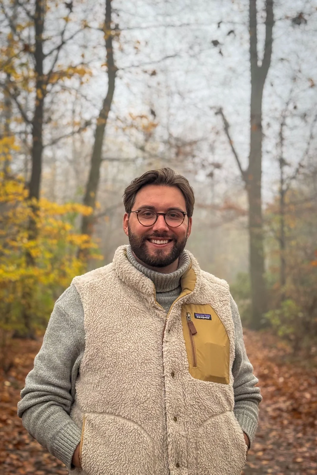Ein lächelnder Mann mit Brille und dunklem Haar, trägt einen hellbeigen Fleece-Weste und einen grauen Pullover, steht in einem herbstlichen Wald mit fallenden Blättern an den Bäumen.