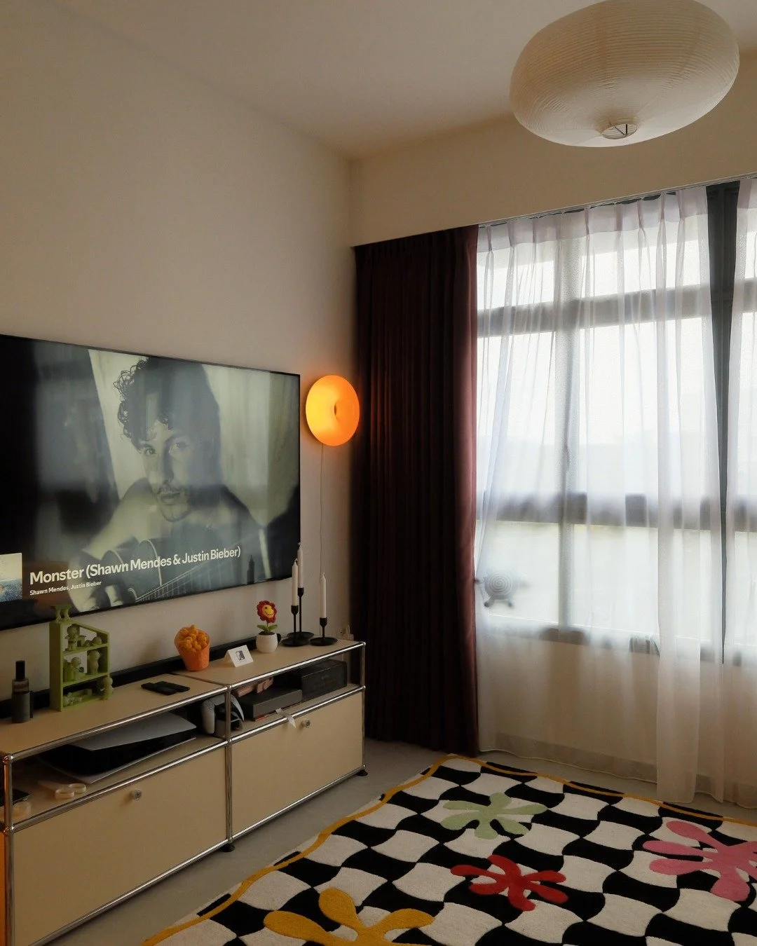 The corner that gets the details right.

A metal frame console sits beneath a large screen, layered with small objects that tell you exactly who lives here. The orb wall sconce glows warm against the curtain, and the rice paper pendant above ties the