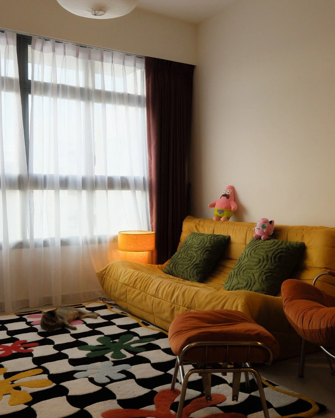 Colour as a way of living.

A mustard yellow sofa sits low and generous, the kind you sink into and don't leave. The graphic rug anchors the whole room in bold black and white with bursts of colour, and the plushies on the sofa backrest make it clear