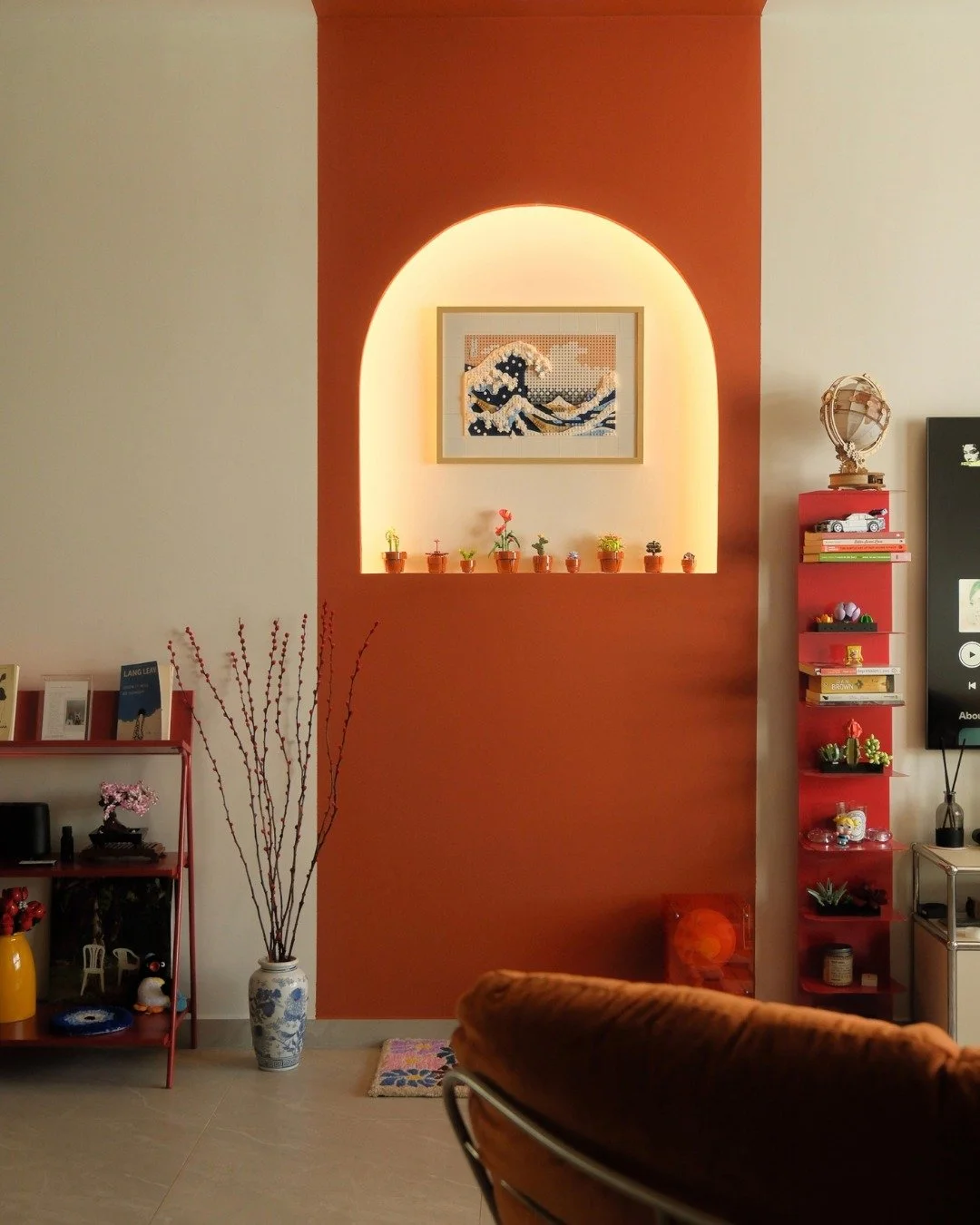 Every shelf has a story.

The orange accent wall anchors the living room with a lit arched niche that frames a Lego art piece of The Great Wave, surrounded by a row of tiny potted plants. Red shelves climb the wall on both sides, packed with collecti