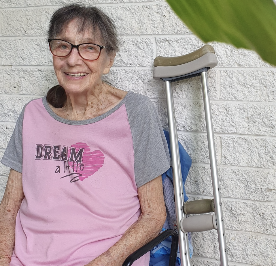An elderly woman with glasses smiling, sitting next to a white brick wall. She is wearing a pink and gray T-shirt with the words 'Dream Big' on it, and has a cane leaning against the wall beside her.