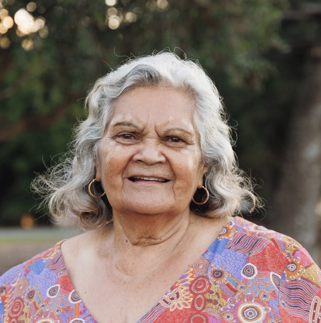 Smiling elderly woman with gray curly hair wearing colorful patterned blouse and hoop earrings outdoors, with trees in the background.